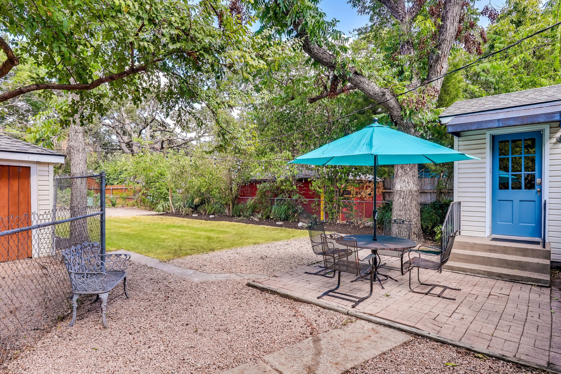 Backyard patio with a turquoise umbrella, metal seating, gravel, and a small lawn beneath mature trees.