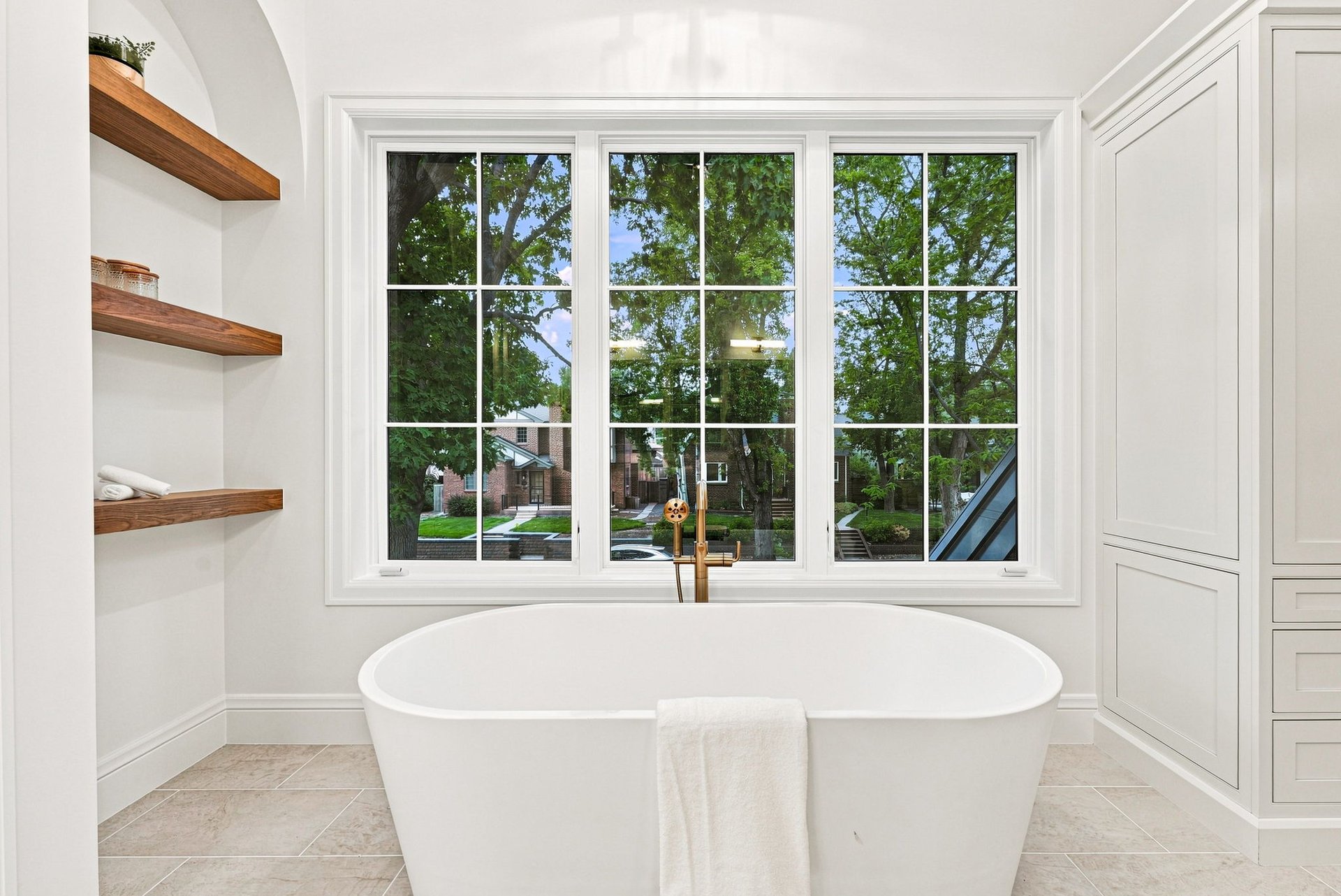 Bathroom with a freestanding soaking tub beneath a large window and open wood shelves.
