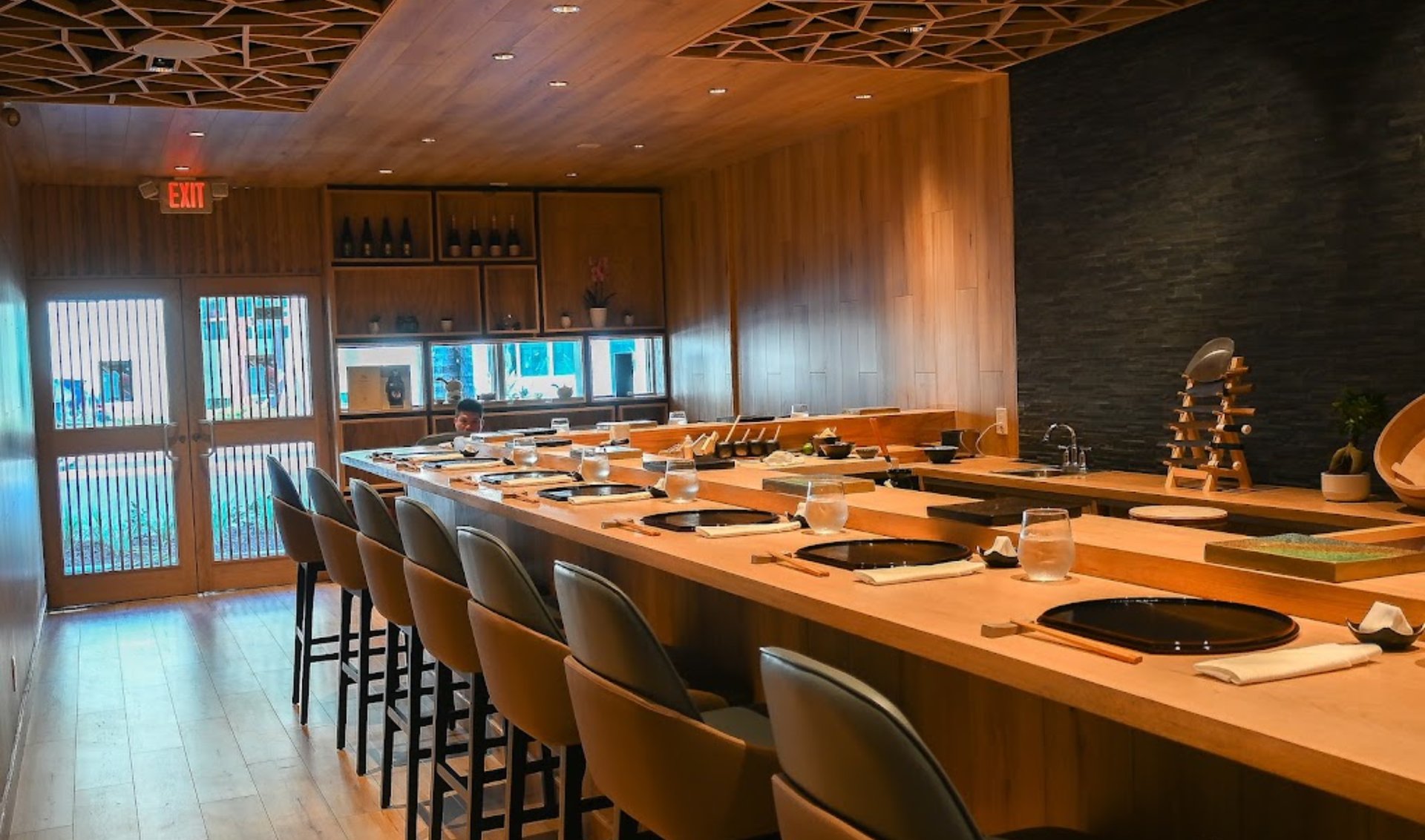 Sushi restaurant counter with wooden seating, individual place settings, and geometric ceiling.