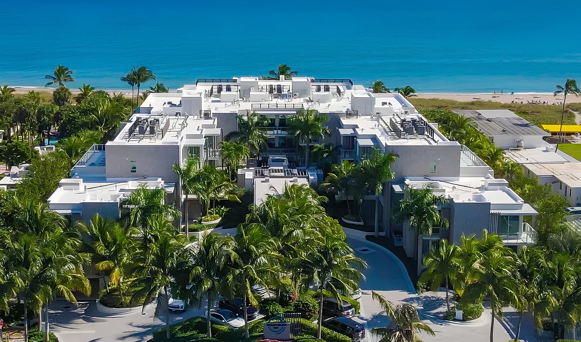 Low-rise beachfront condominium with rooftop terraces, a central pool, palm trees, and ocean views.