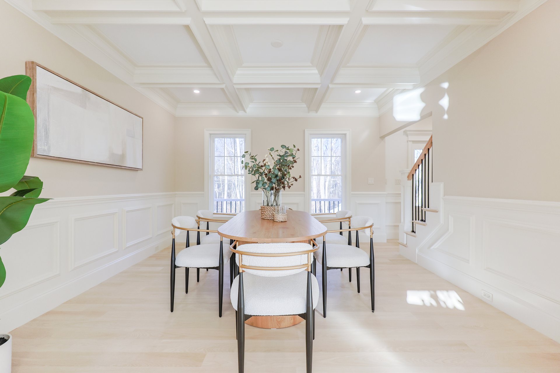 Dining room with an oval wood table, white chairs, coffered ceiling, and staircase.