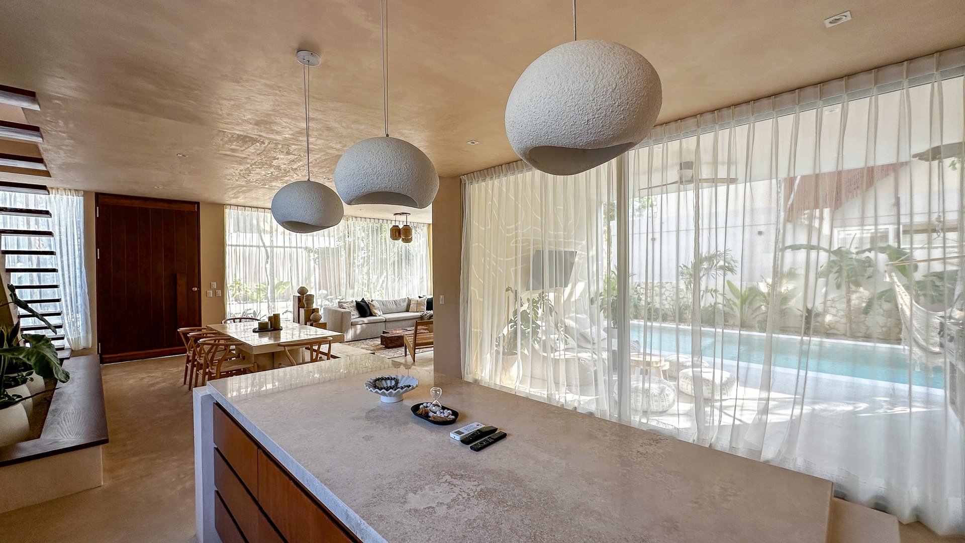 Open-plan kitchen and dining area with sculptural pendant lights and a pool visible through sheer curtains.