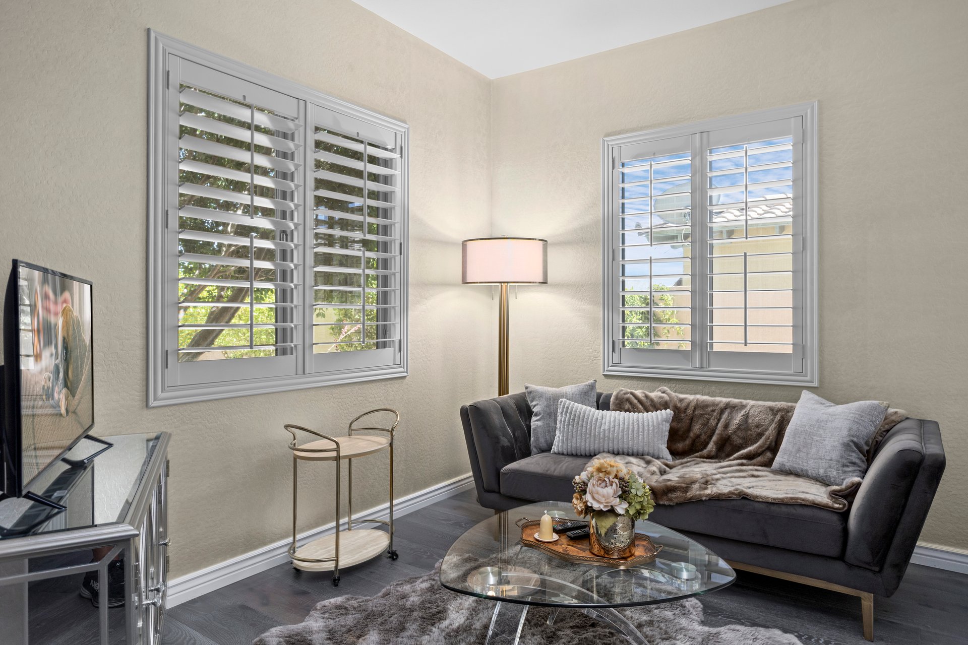 Staged living room corner with a dark sofa, glass coffee table, floor lamp, TV stand, and two windows with white shutters.