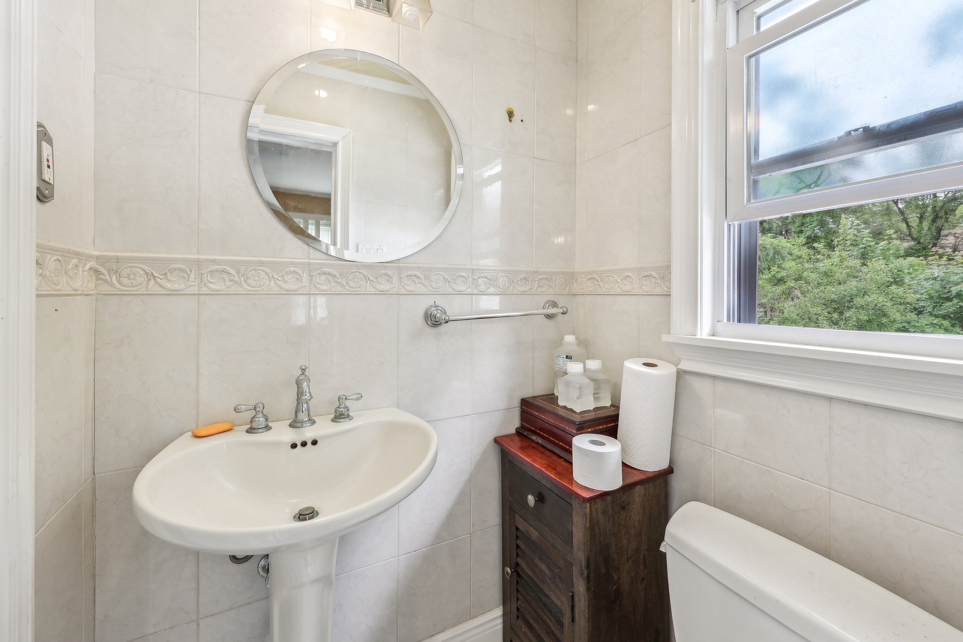 Bathroom with pedestal sink, round mirror, tiled walls, and a window overlooking trees.