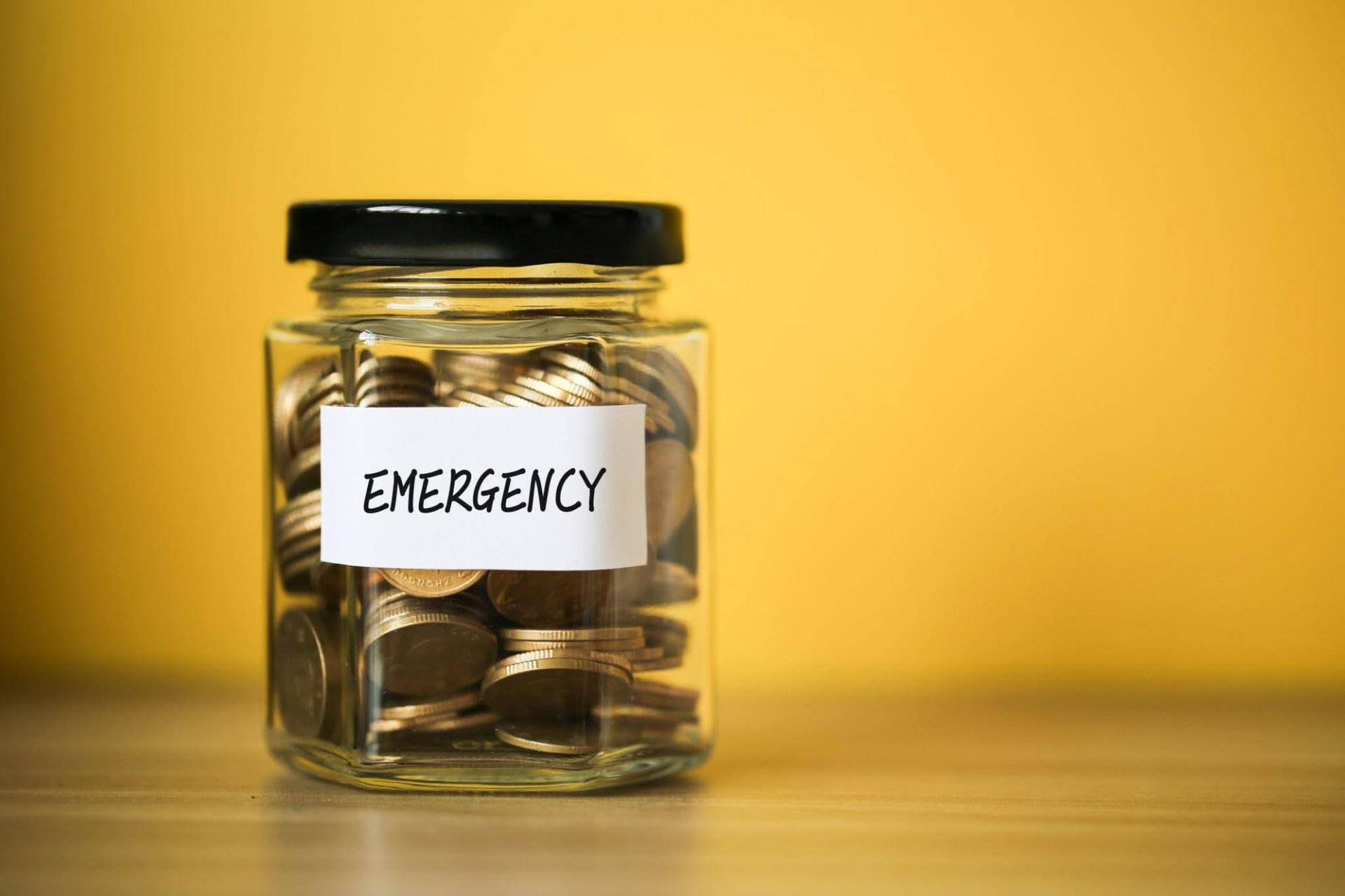 a-glass-jar-filled-with-coins-labeled-emergency-sits-on-a-wooden-surface-against-a-bright-yellow-background