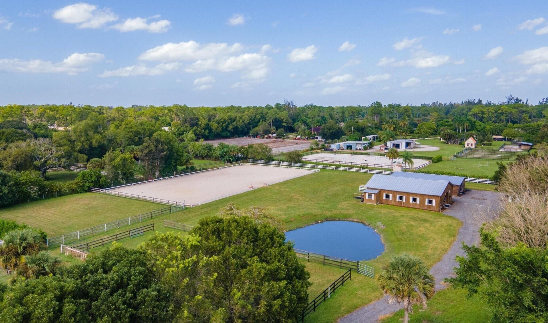 Aerial view of an equestrian property with fenced riding arenas, paddocks, a barn, and a pond.