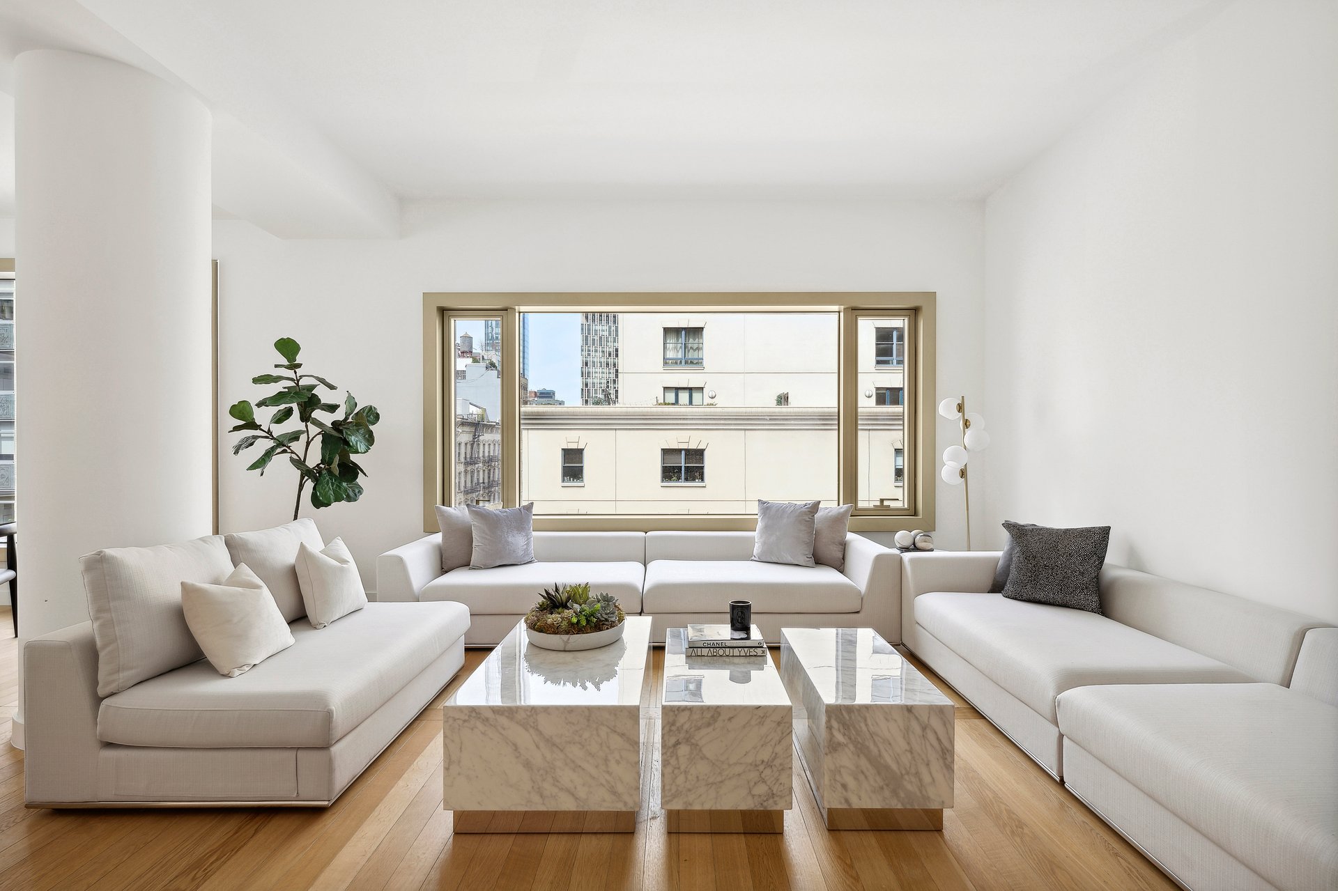 Bright living room with white sectional sofas, marble coffee tables, and a broad city-facing window.
