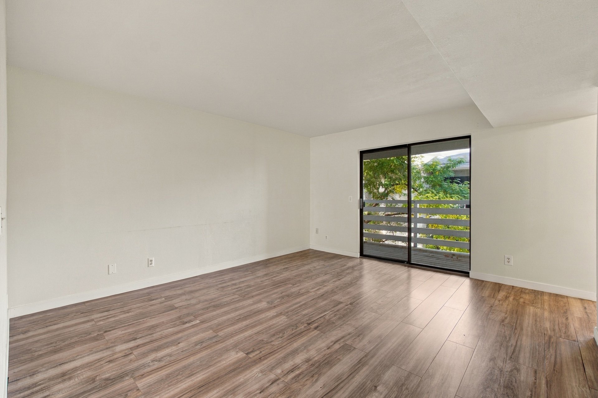 Empty living room with wood-look flooring and a sliding glass door to a tree-lined balcony.