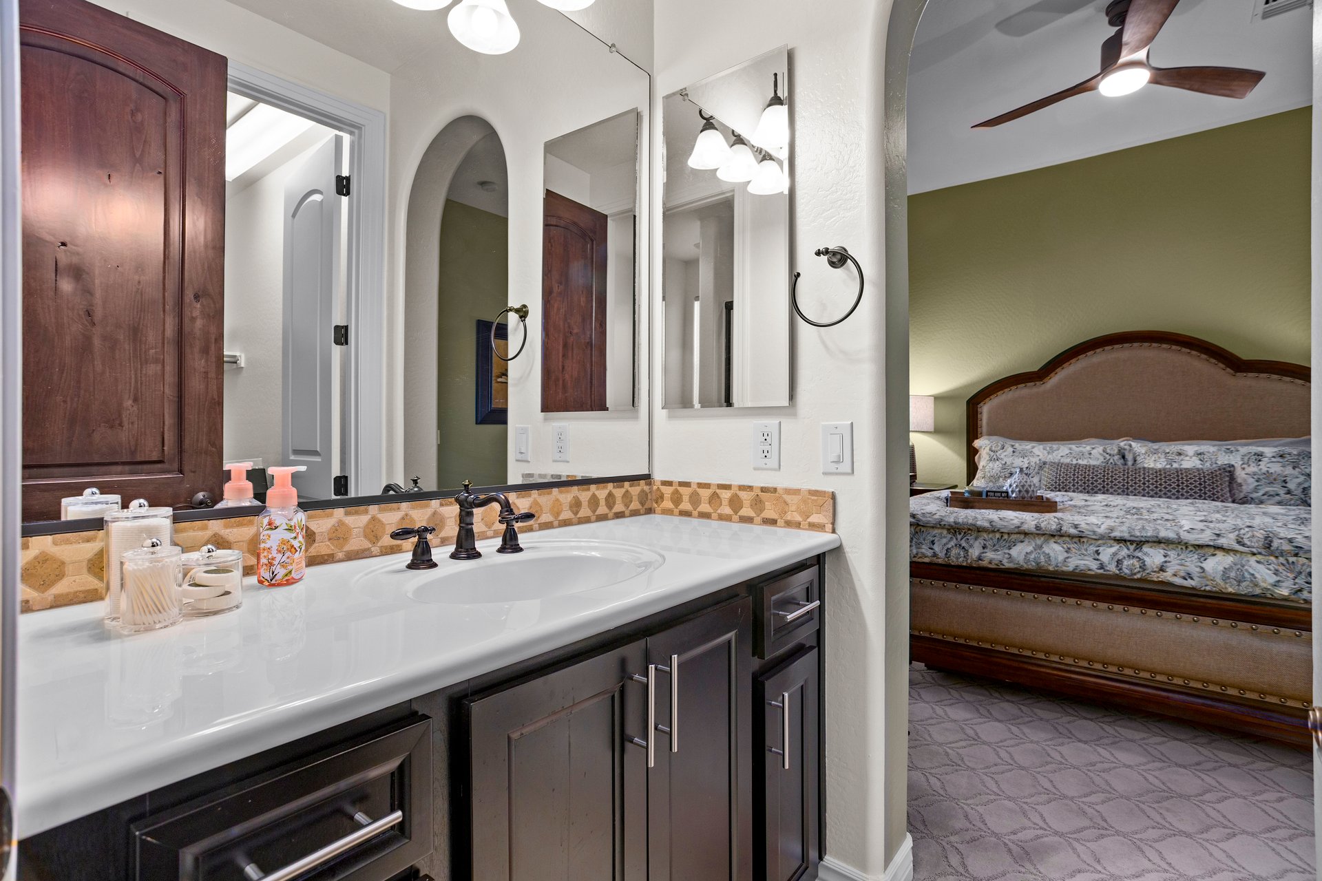 Master bathroom vanity with double mirrors and dark cabinetry, looking through to a bedroom with a bed and ceiling fan.