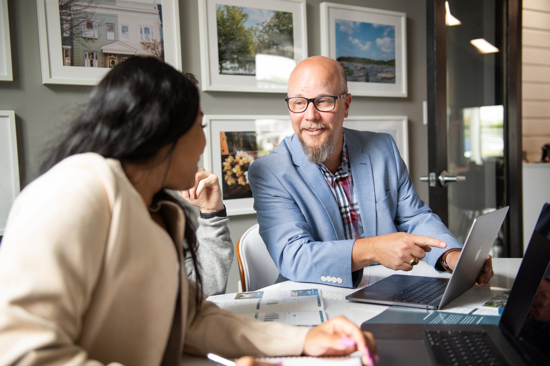A real estate agent points at a laptop while discussing documents with a client.