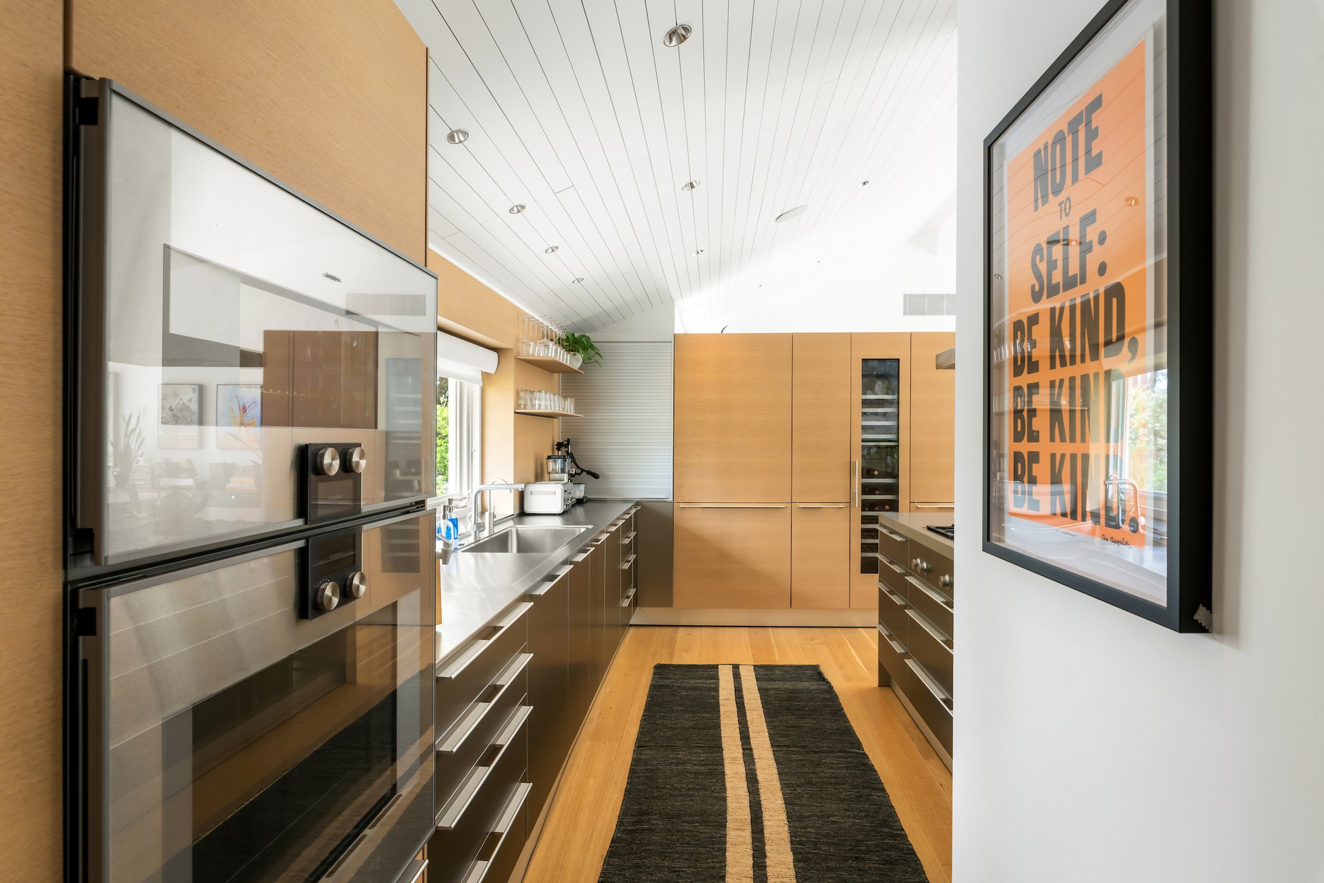 Modern galley kitchen with warm wood cabinetry, stainless appliances, and a framed orange poster.