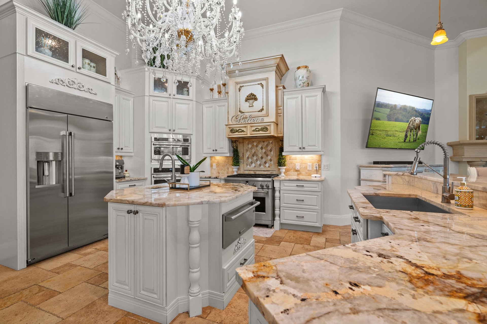 Ornate white kitchen with marble counters, stainless appliances, and a crystal chandelier.
