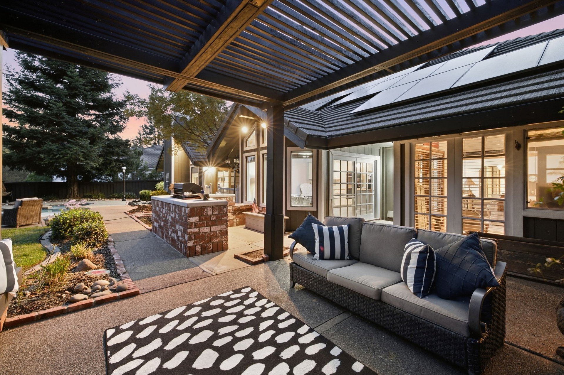 Covered patio with pergola, outdoor sofa, brick grill island, and pool at dusk.