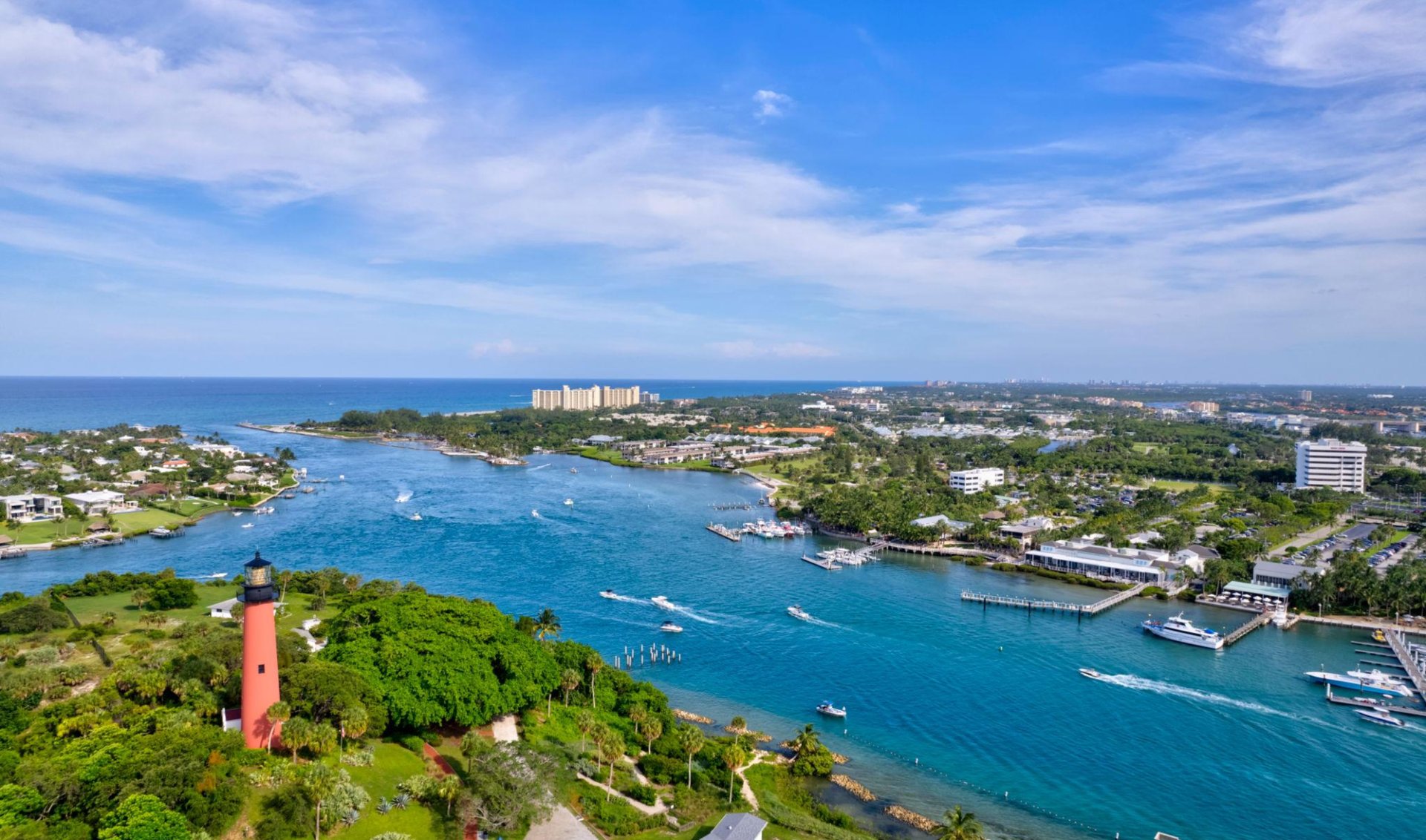 Aerial view of a coastal inlet with boats, waterfront homes, and a red lighthouse.