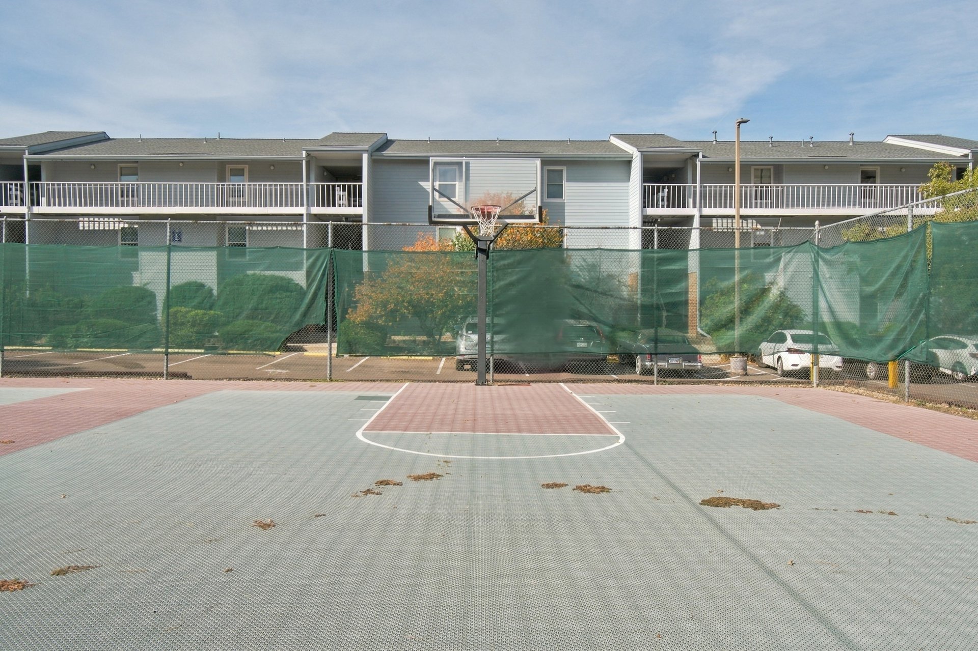 Outdoor basketball court with a hoop, green-screened fence, and apartment building behind it.