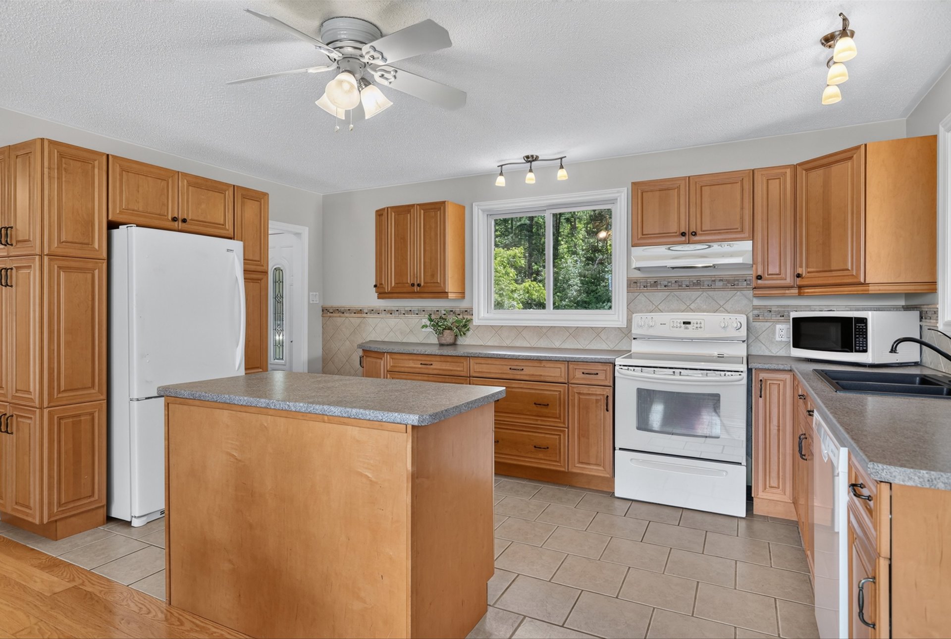 Kitchen with honey-colored wood cabinets, a central island, and a window overlooking leafy trees.