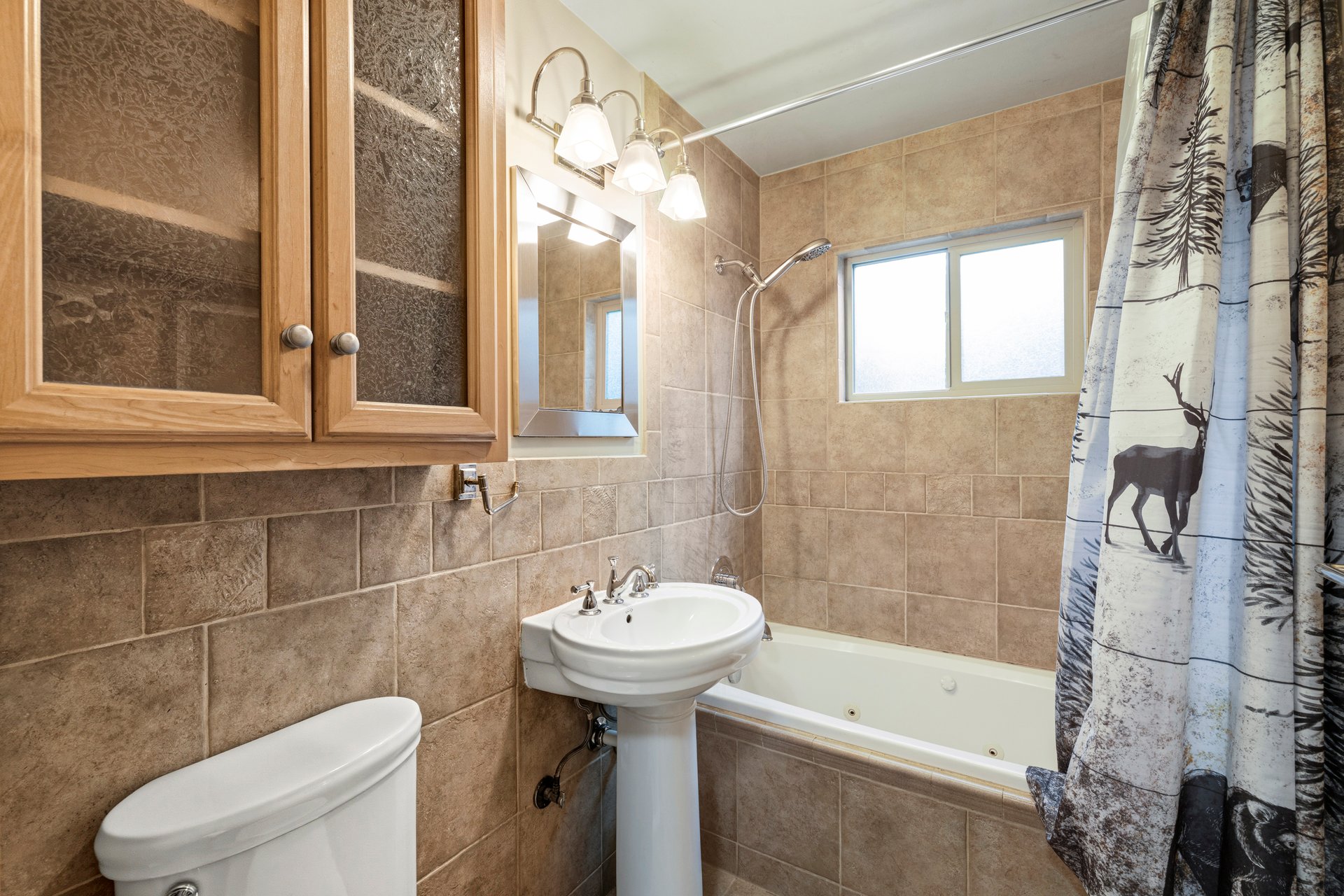 Tiled bathroom with a bathtub, pedestal sink, wooden cabinets, and deer-print shower curtain.