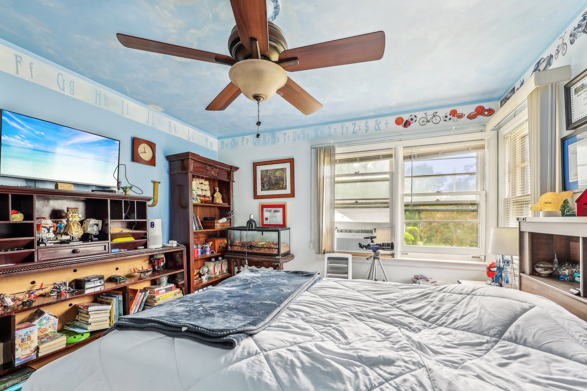 Furnished bedroom with a large bed, dark wood shelving, ceiling fan, and bright windows.
