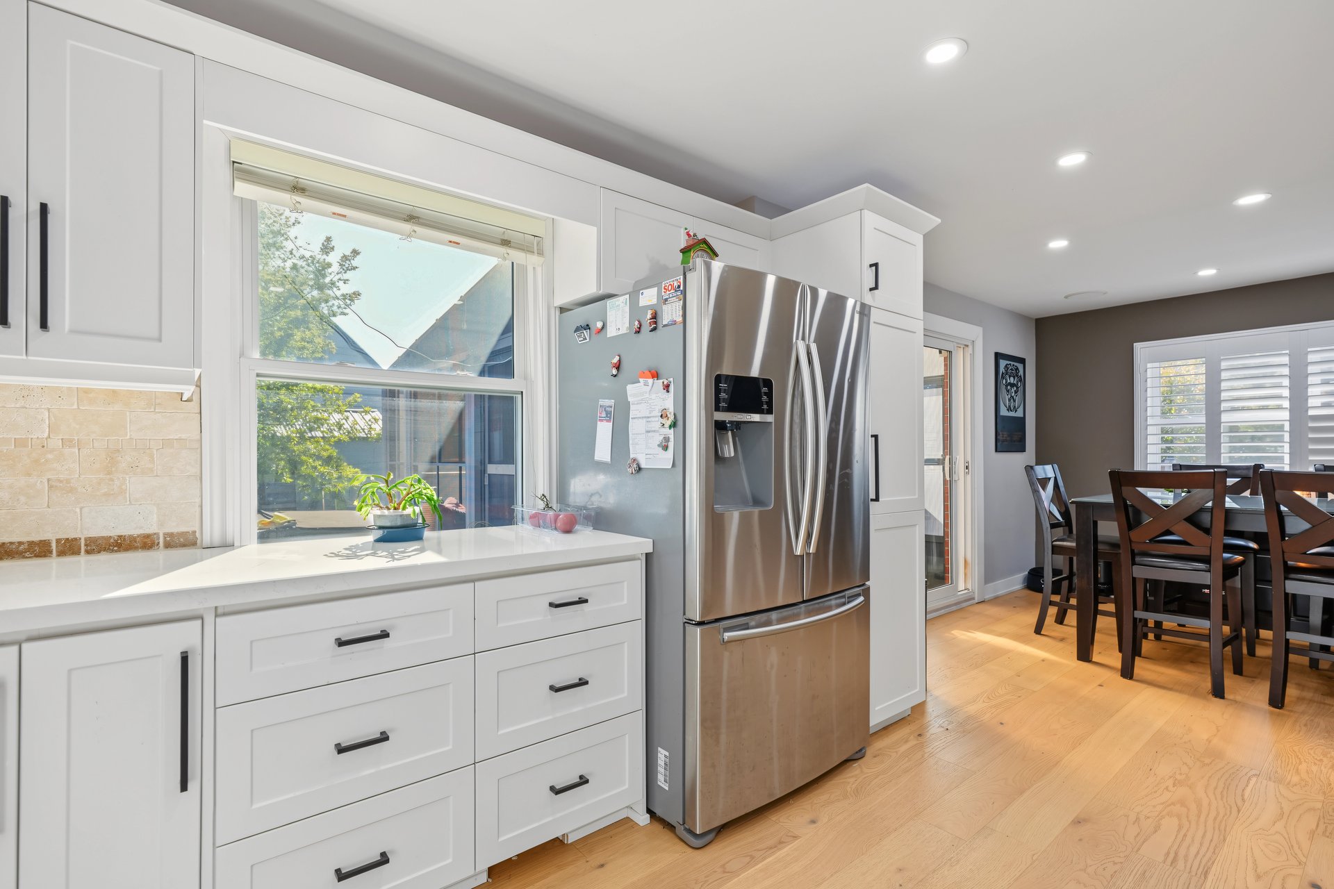 Bright kitchen with white cabinetry, stainless-steel refrigerator, large window, and adjoining dining area.