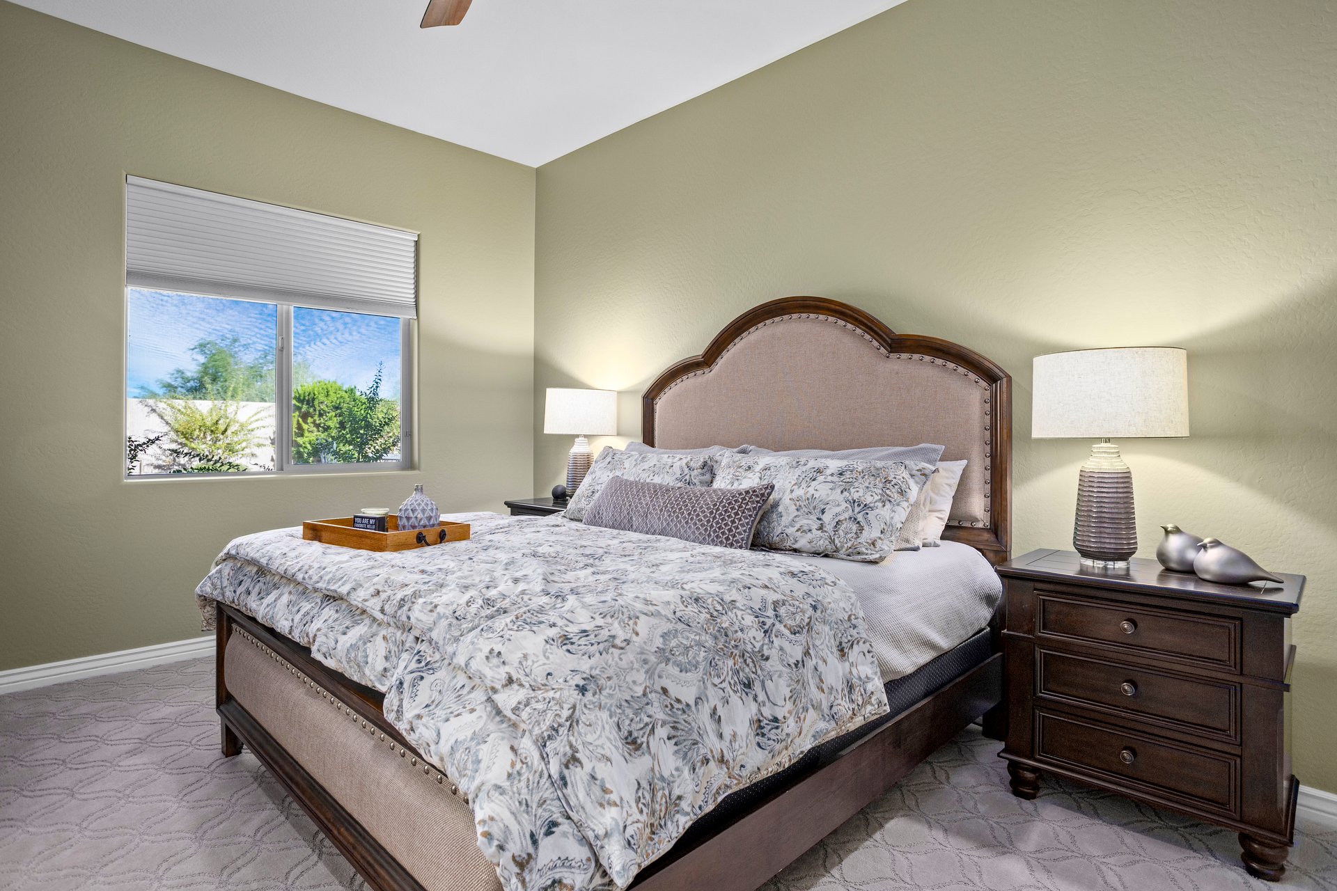 A furnished bedroom with a large bed, matching nightstands and lamps, and a window showing greenery outside.
