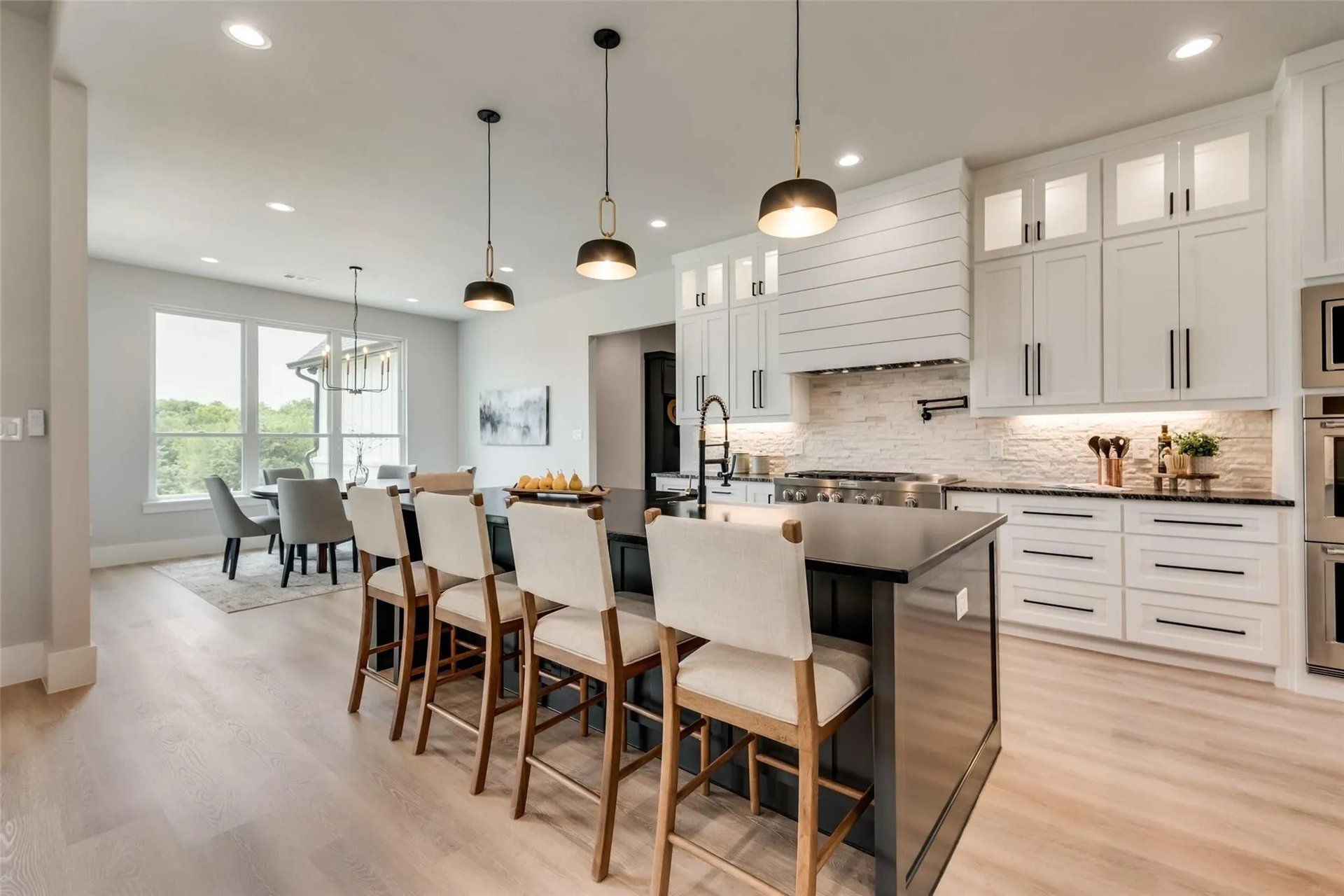 Modern white kitchen with a large dark island and dining area beside windows overlooking trees.