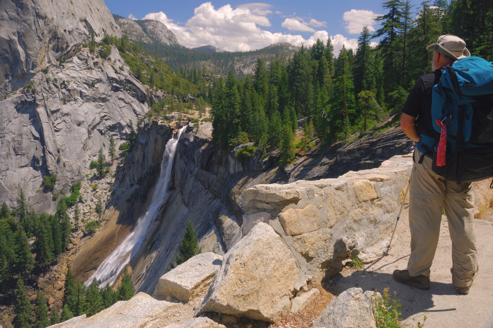 Hiker overlooks a Yosemite waterfall and forested granite cliffs from a rocky trail.