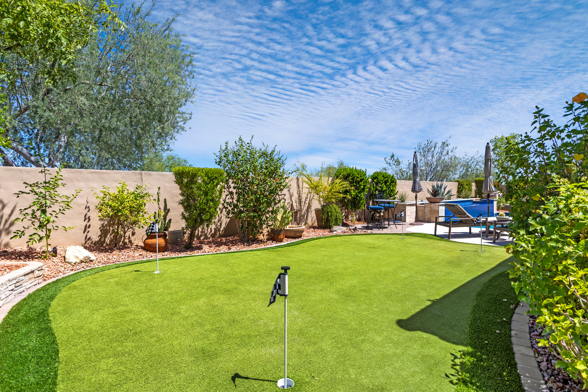 Landscaped backyard with artificial turf putting green, patio seating, and a pool or spa area under a blue sky.