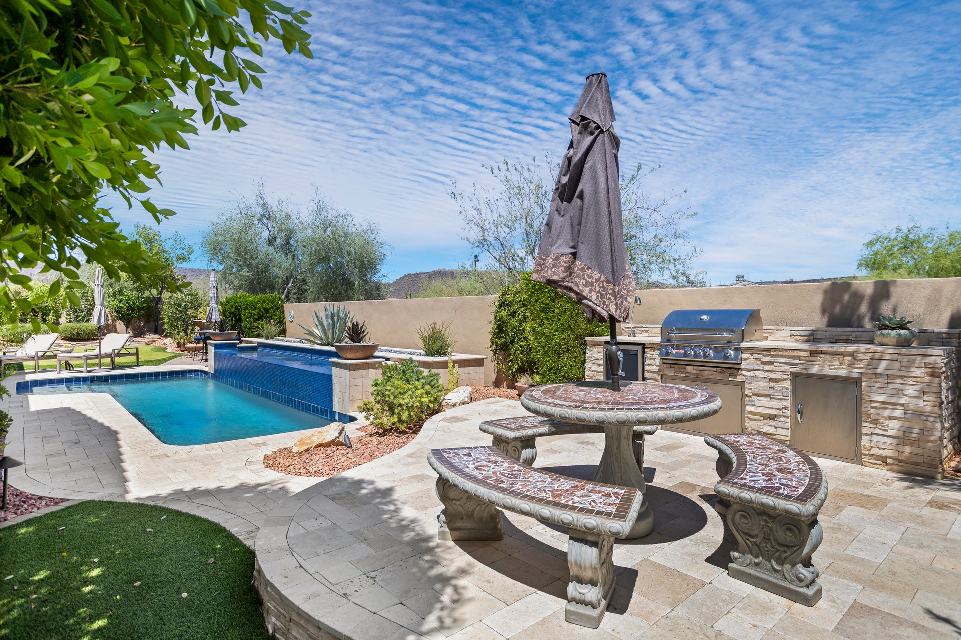 A landscaped backyard patio with a swimming pool, stone dining table and benches, an umbrella, and an outdoor grill under a bright blue sky.