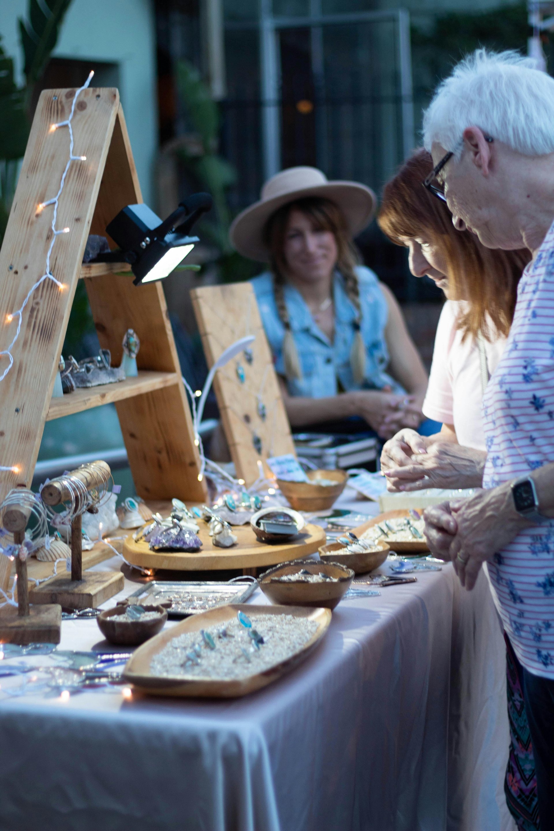 People browsing a jewelry display at an outdoor market, with various handmade pieces showcased on wooden tables.