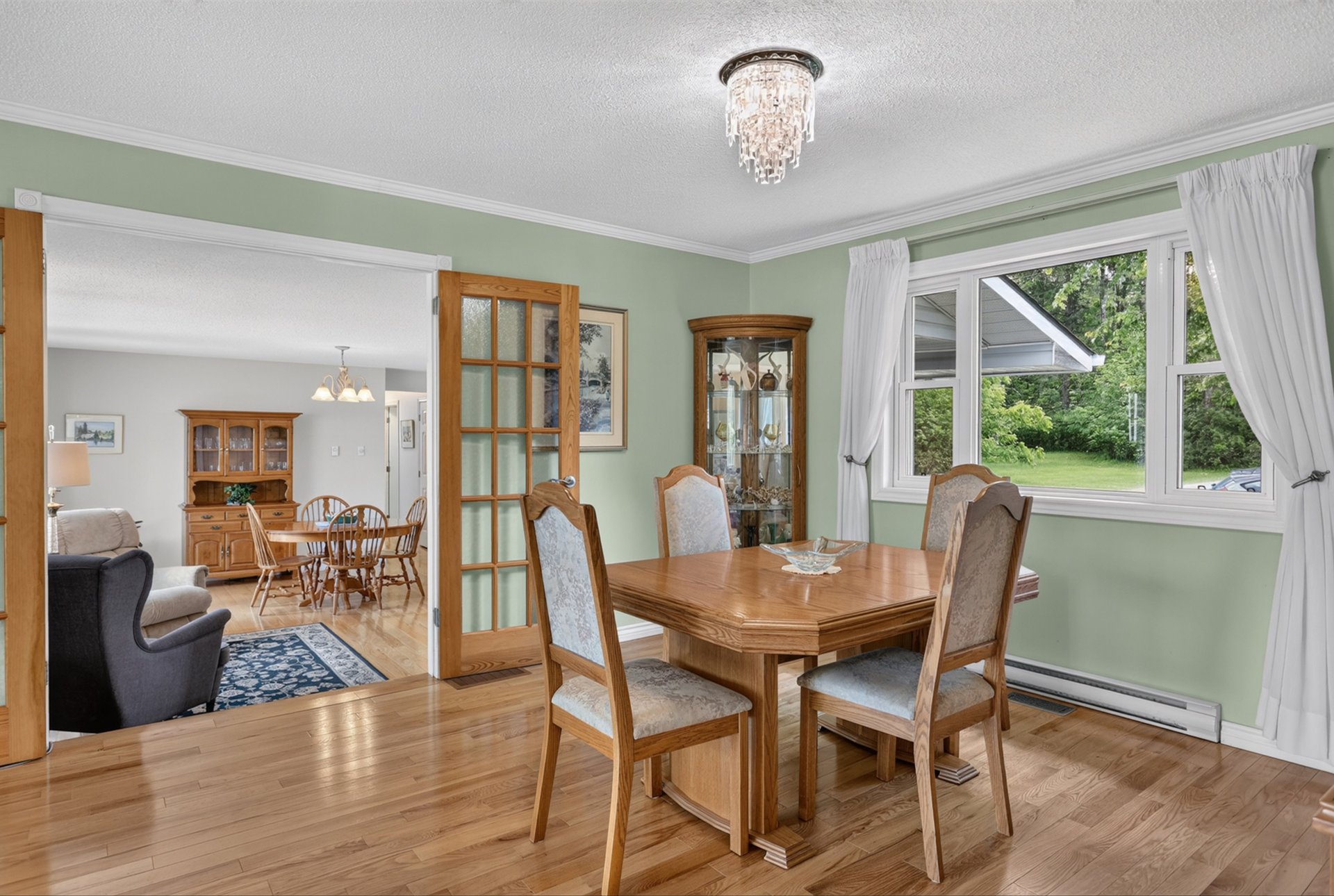 Dining room with a wooden table, pale green walls, hardwood floors, and large curtained windows.