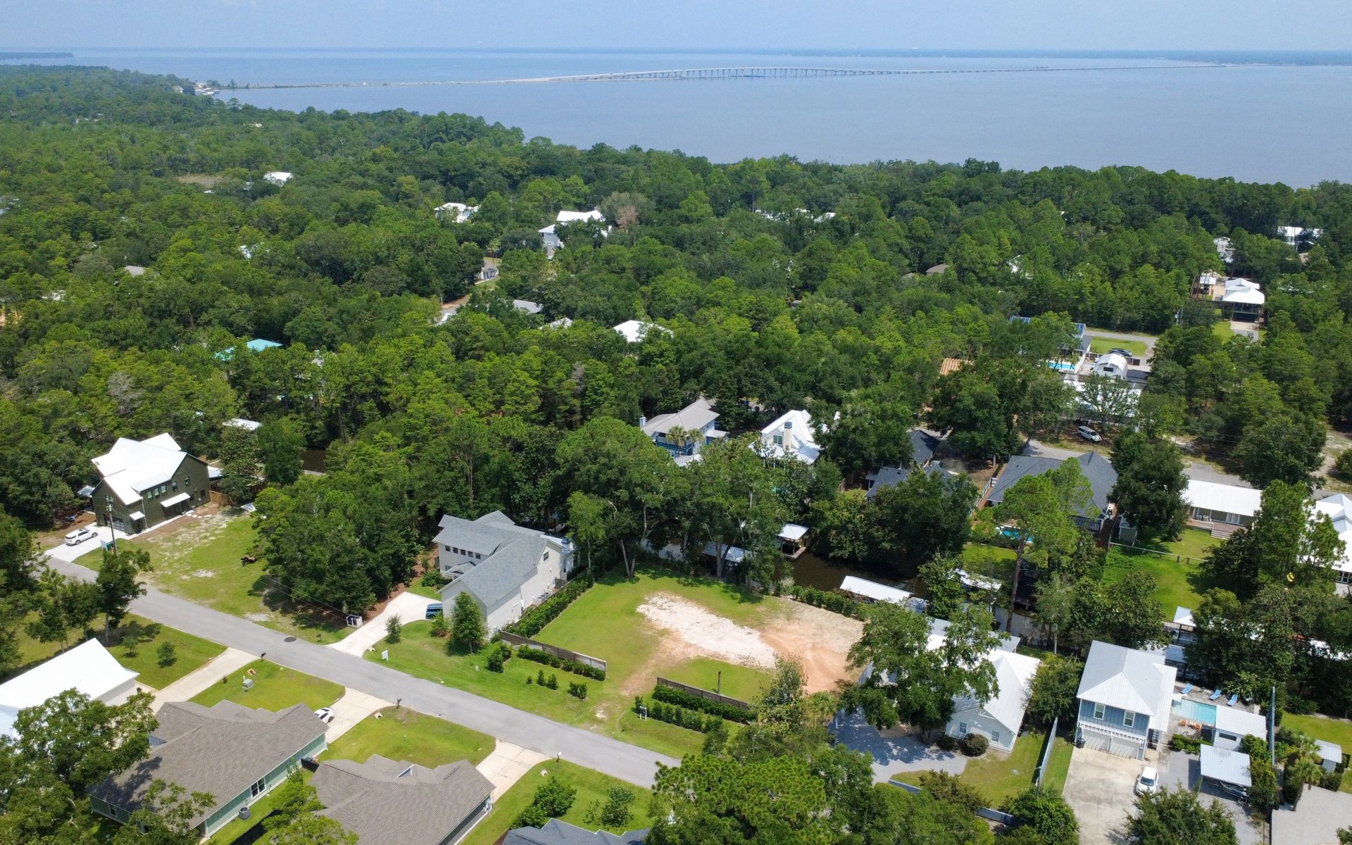 Aerial neighborhood beside a bay, with dense trees, detached homes, and a long bridge across the water.