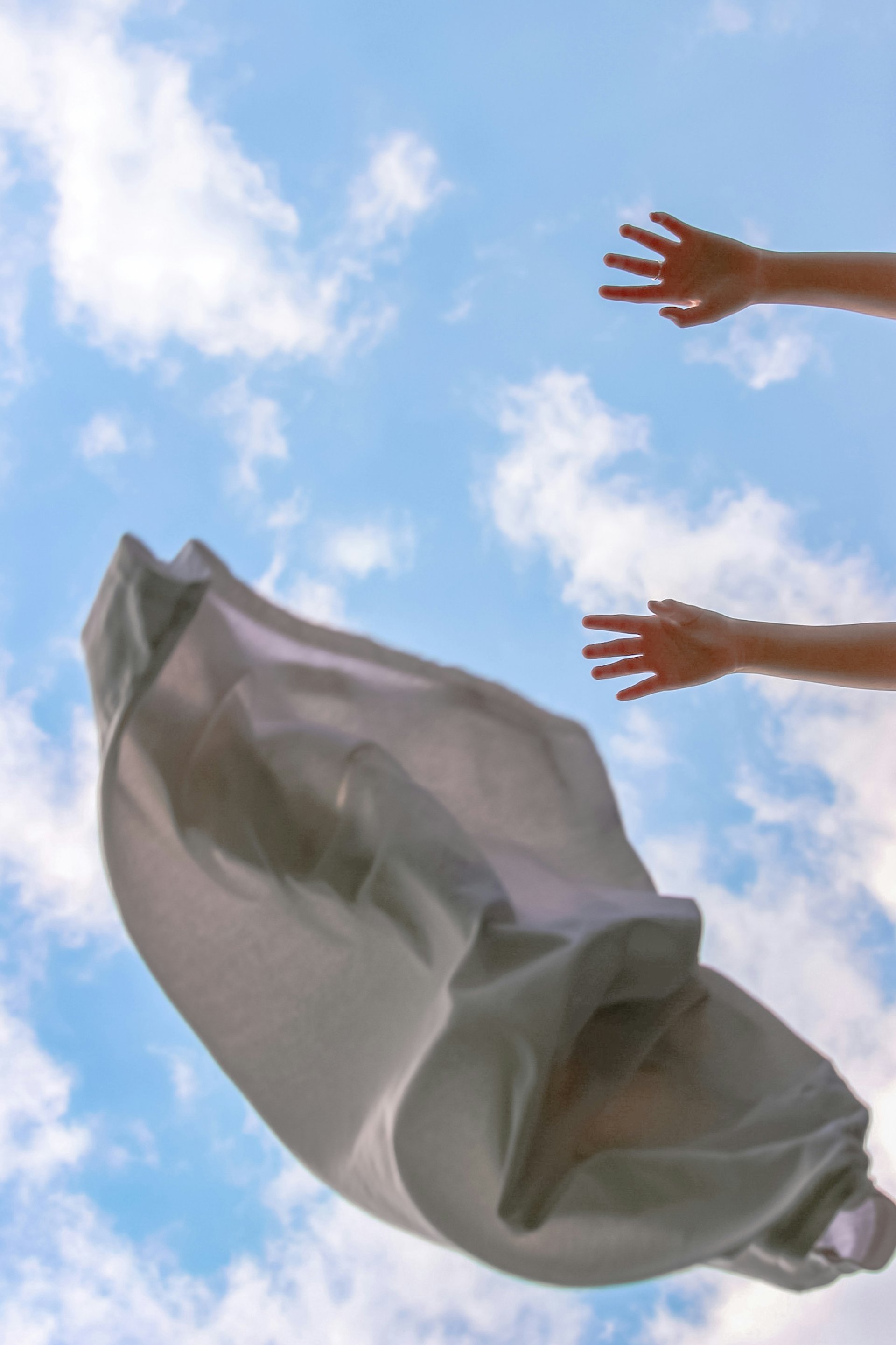 A light garment billows in the breeze against a blue sky as two hands reach toward it.