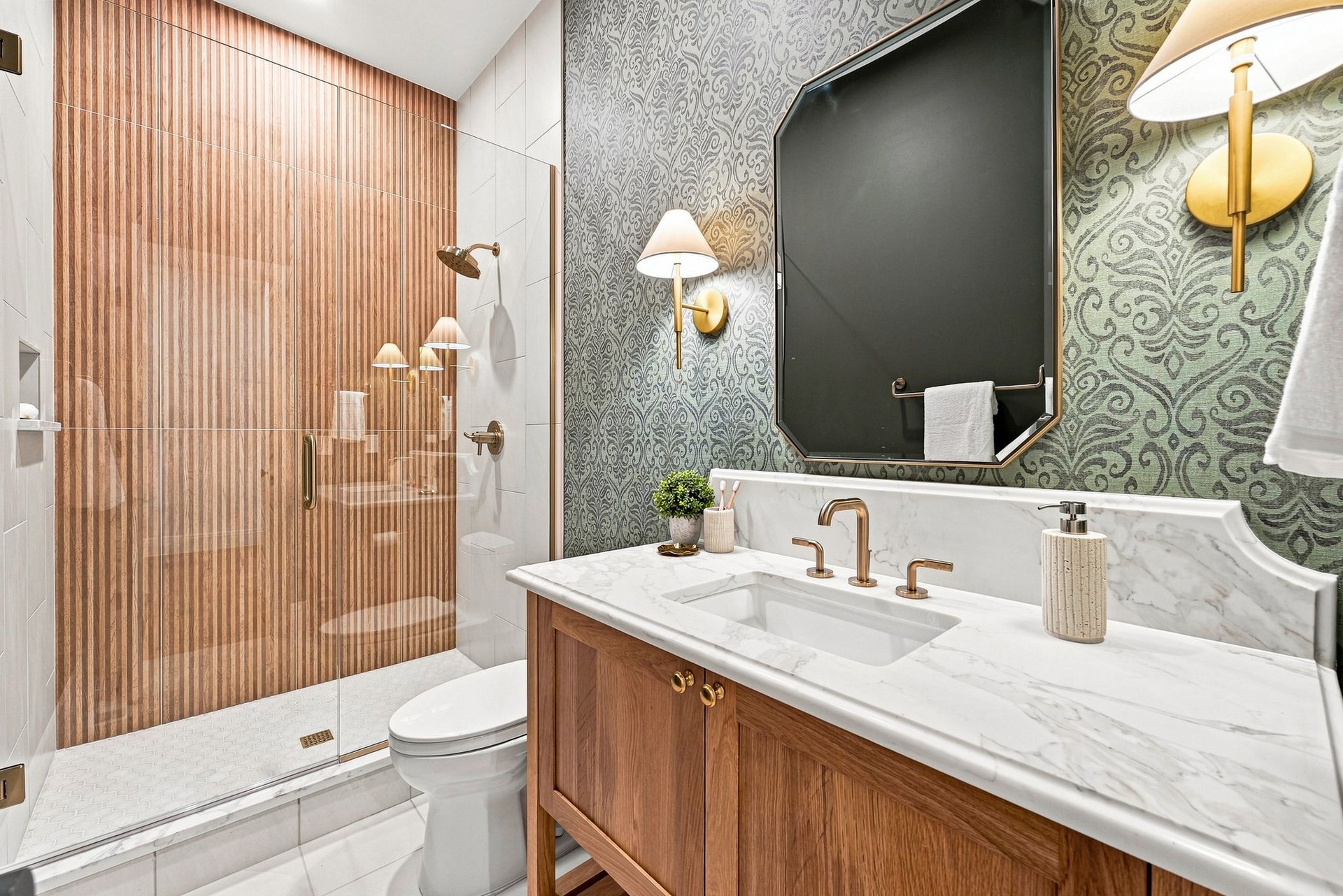 Bathroom with a wood-look tiled shower, marble vanity, patterned wallpaper, and dark-framed mirror.