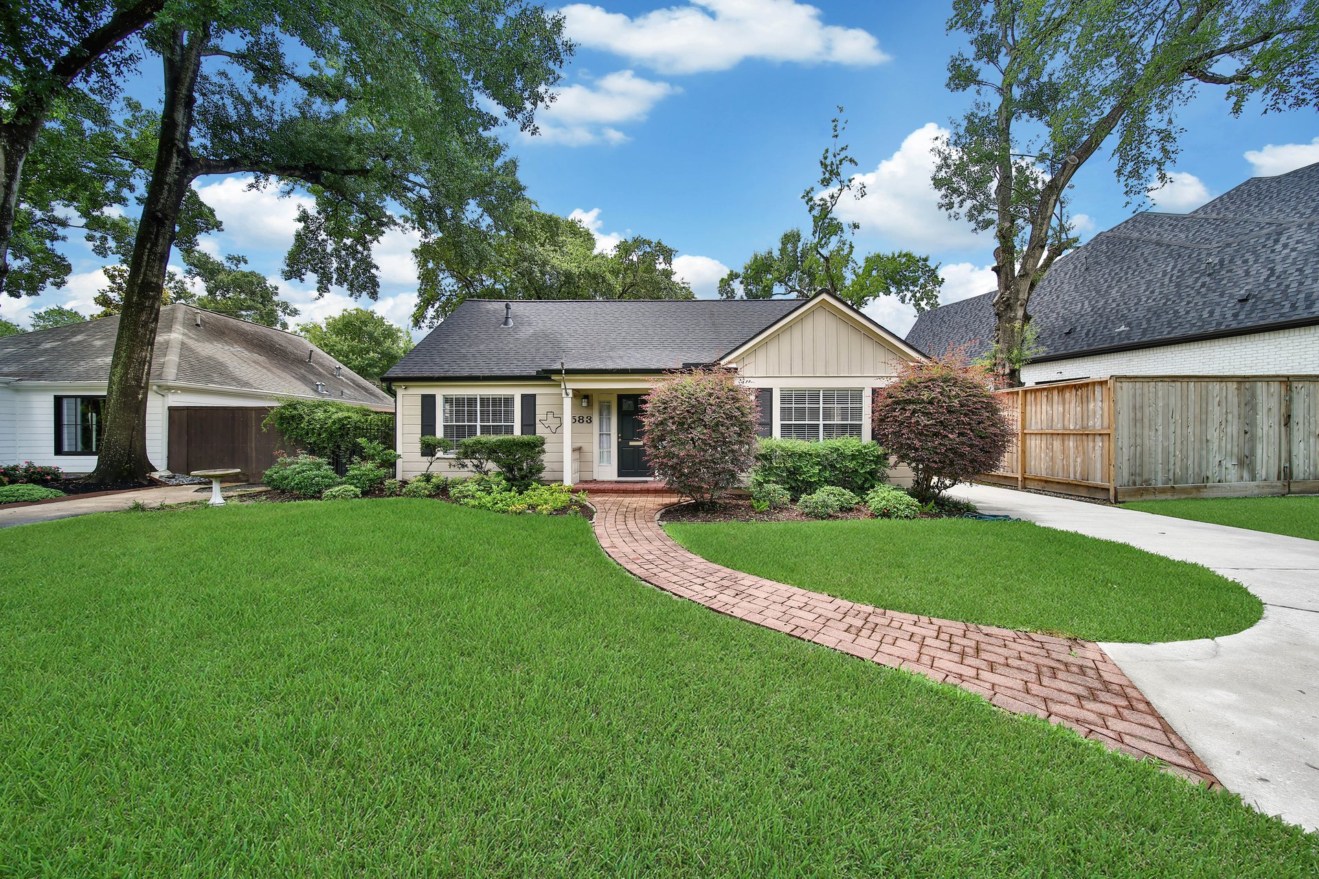 Single-story beige house with a curved brick walkway, landscaped shrubs, and a broad green lawn.