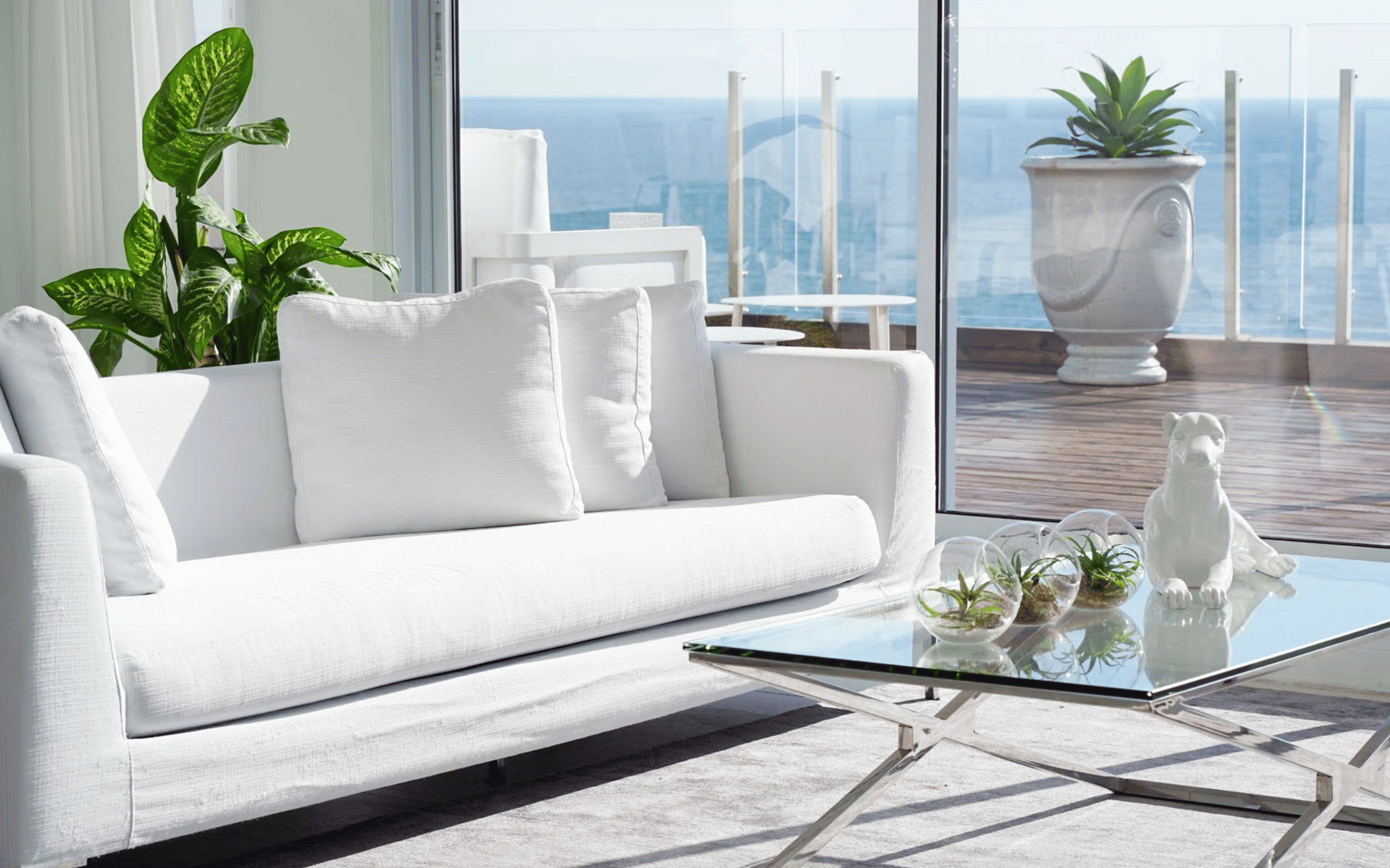 Oceanfront living room with a white sofa, glass coffee table, and sunlit deck view.