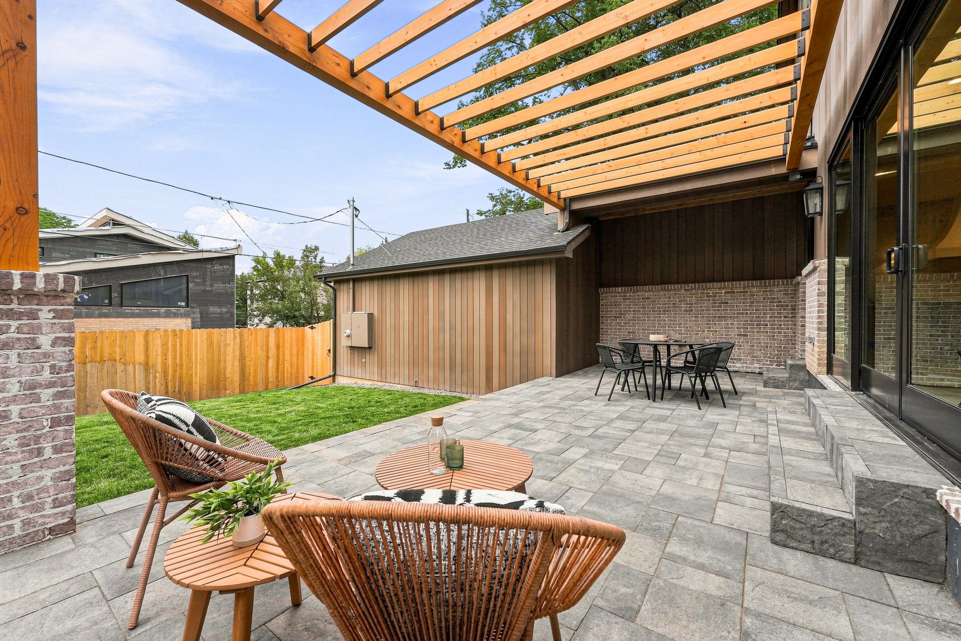 Stone patio with wicker seating and dining table beneath a timber pergola, beside a small fenced lawn.