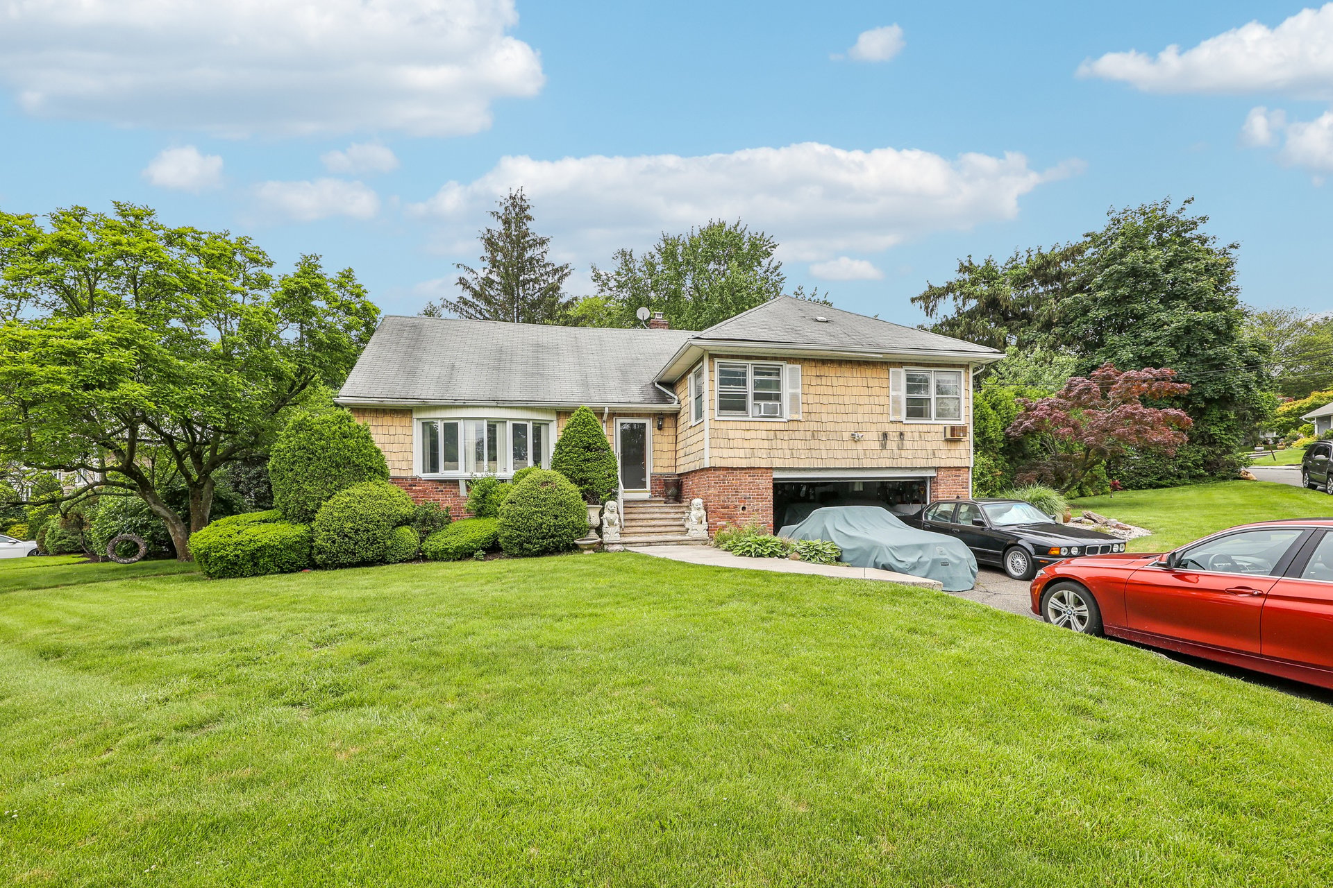 Two-story shingle-and-brick house with a broad lawn, landscaped shrubs, and cars in the driveway.