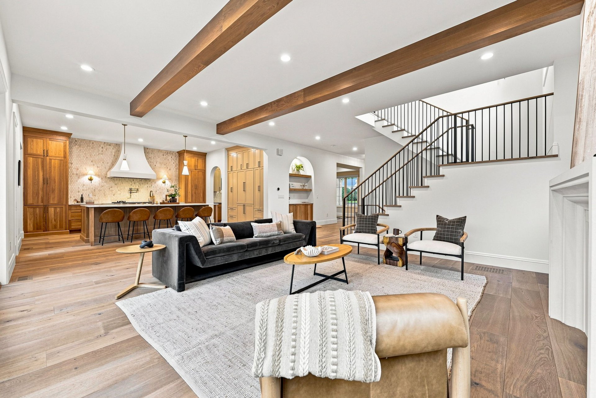 Open-plan living room with exposed wood beams, adjacent kitchen, and curved staircase.