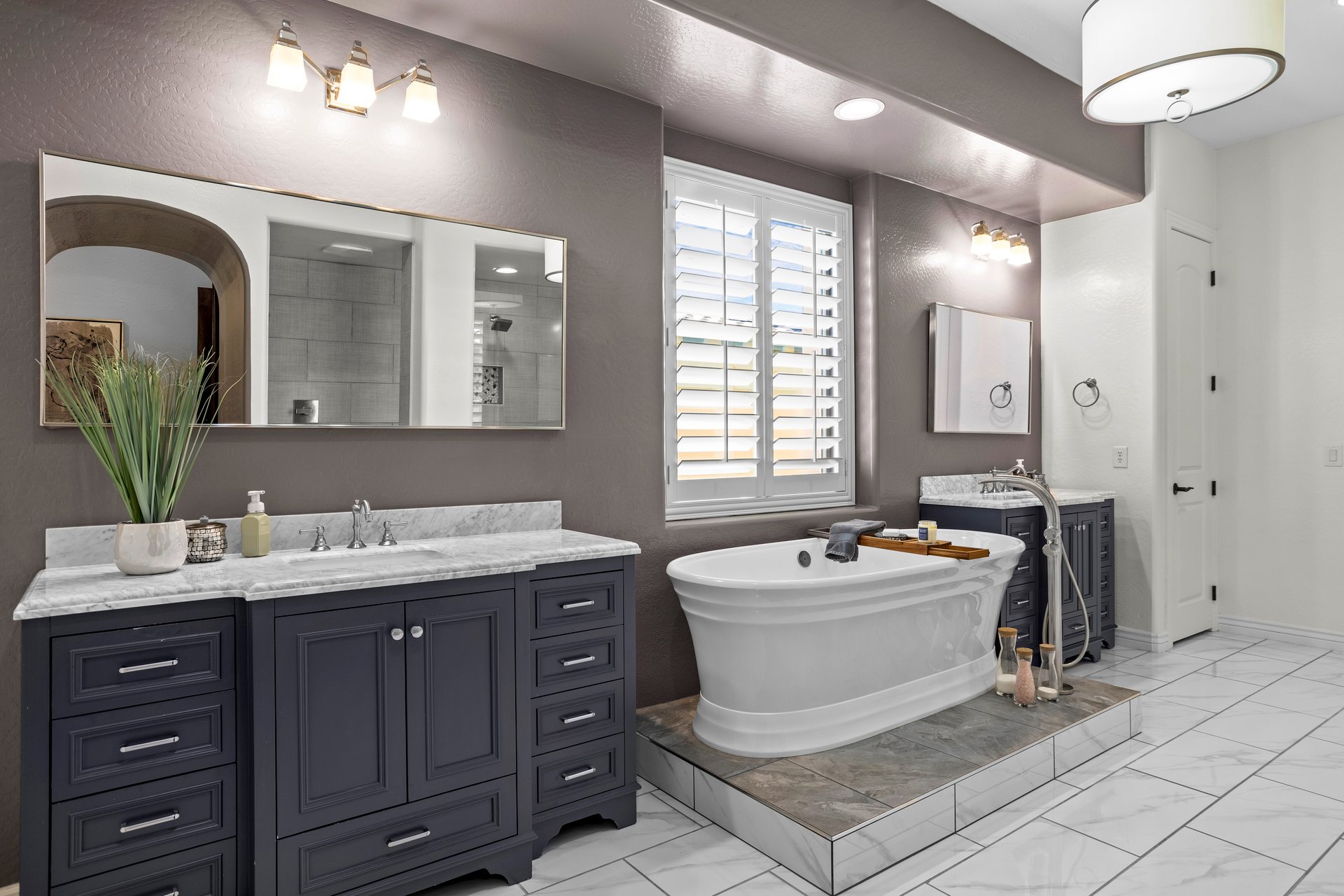 Spacious primary bathroom with dark dual-sink vanity, large mirrors, freestanding soaking tub on a tiled platform, and window shutters.