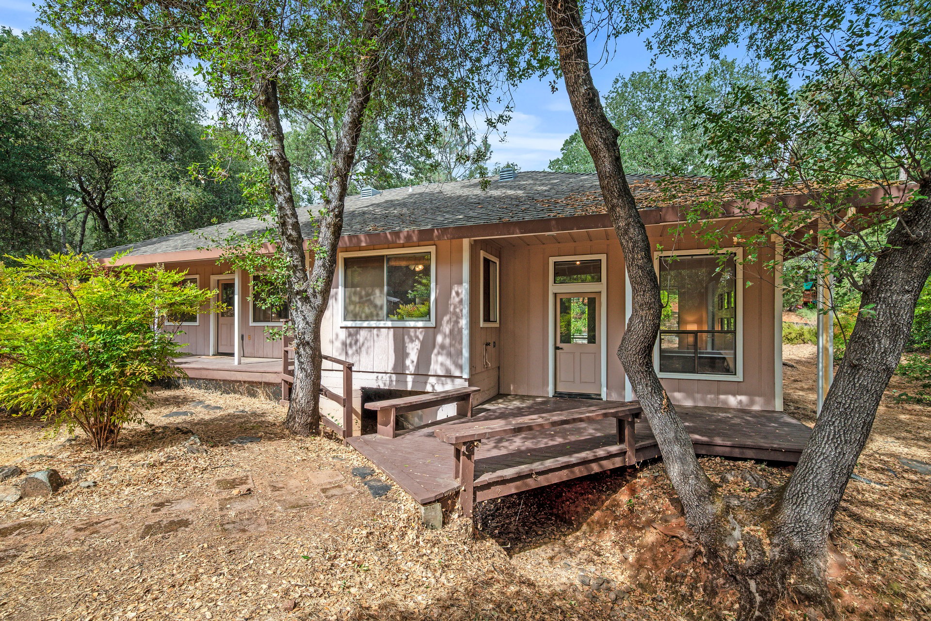 Single-story home with a raised wooden deck, framed by mature trees and shrubs.