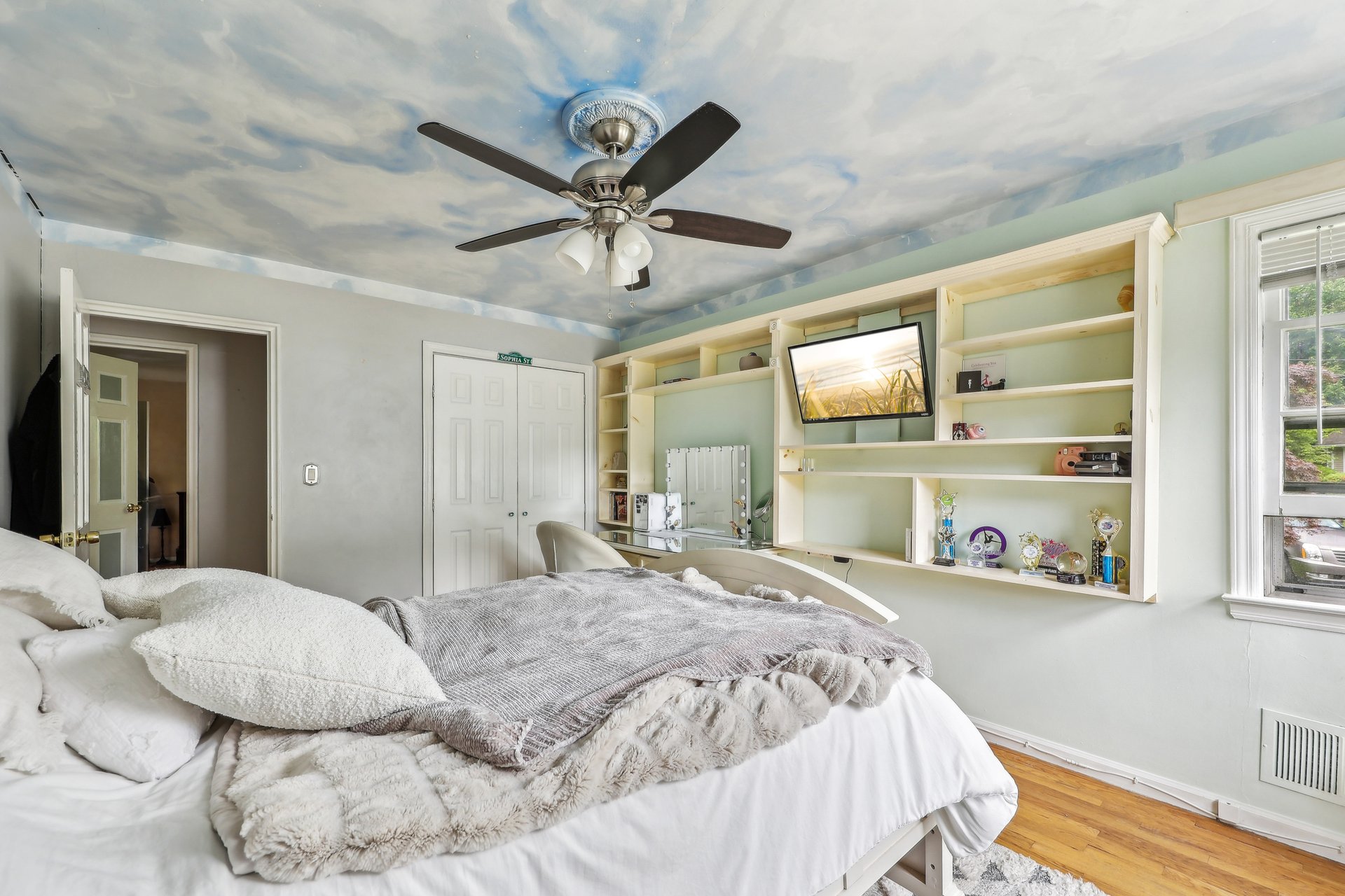 Bedroom with a large bed, built-in shelves, ceiling fan, and cloudy sky ceiling mural.