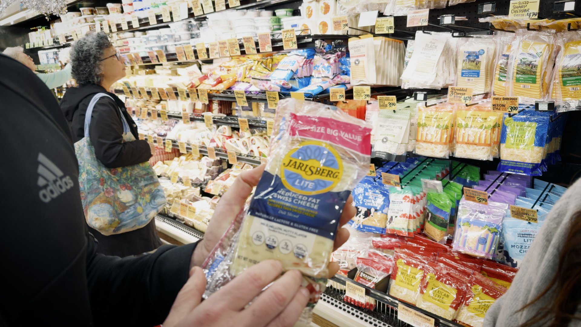 A person holds a package of Jarlsberg Lite cheese in a grocery store aisle filled with various cheese products.