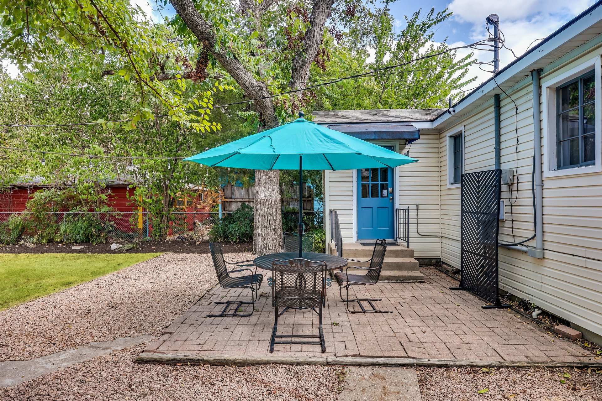 Backyard patio with metal seating under a turquoise umbrella beside a house with a bright blue door.