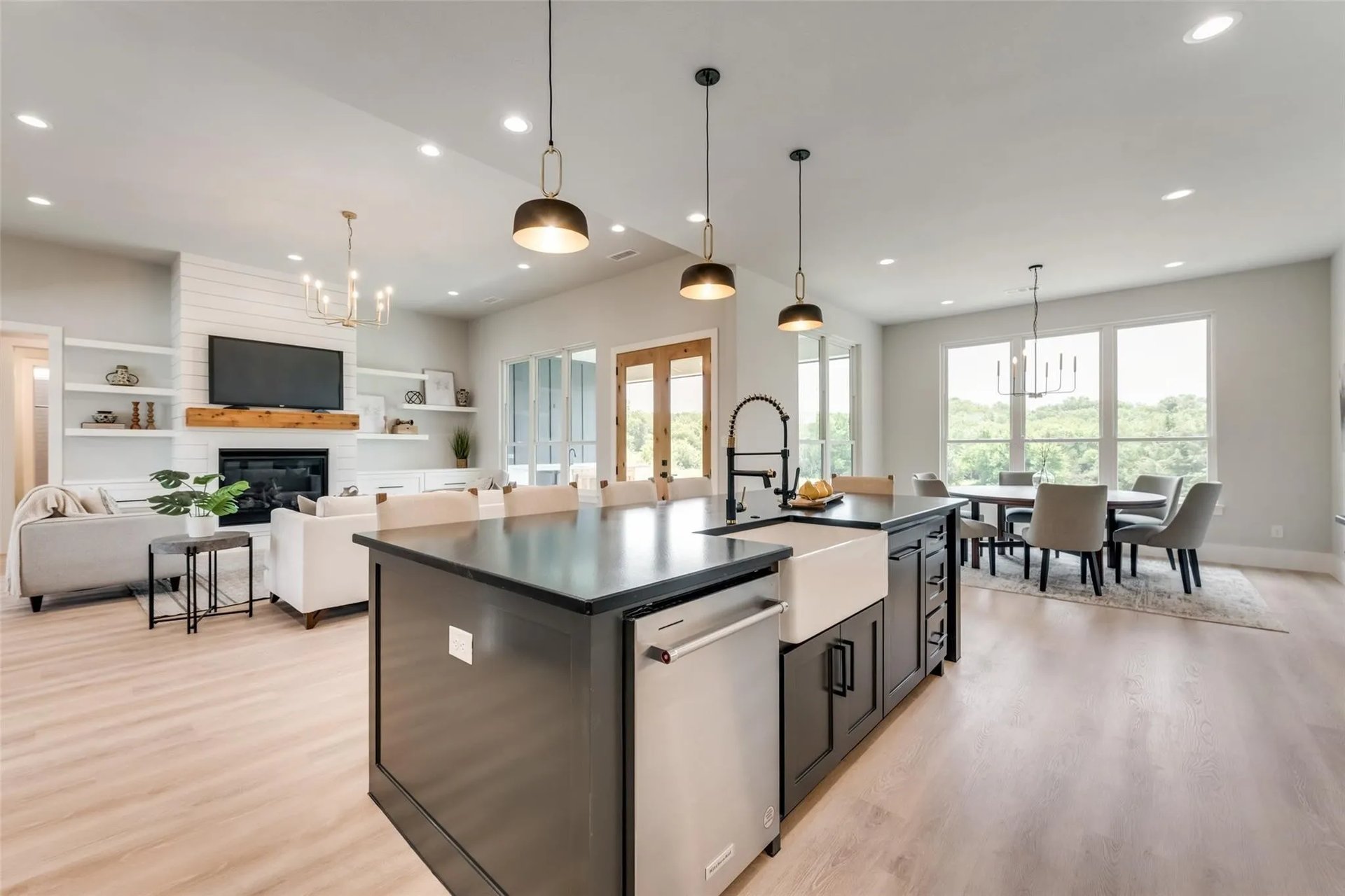 Open-plan kitchen with a large dark island, adjoining living and dining areas, and windows overlooking trees.