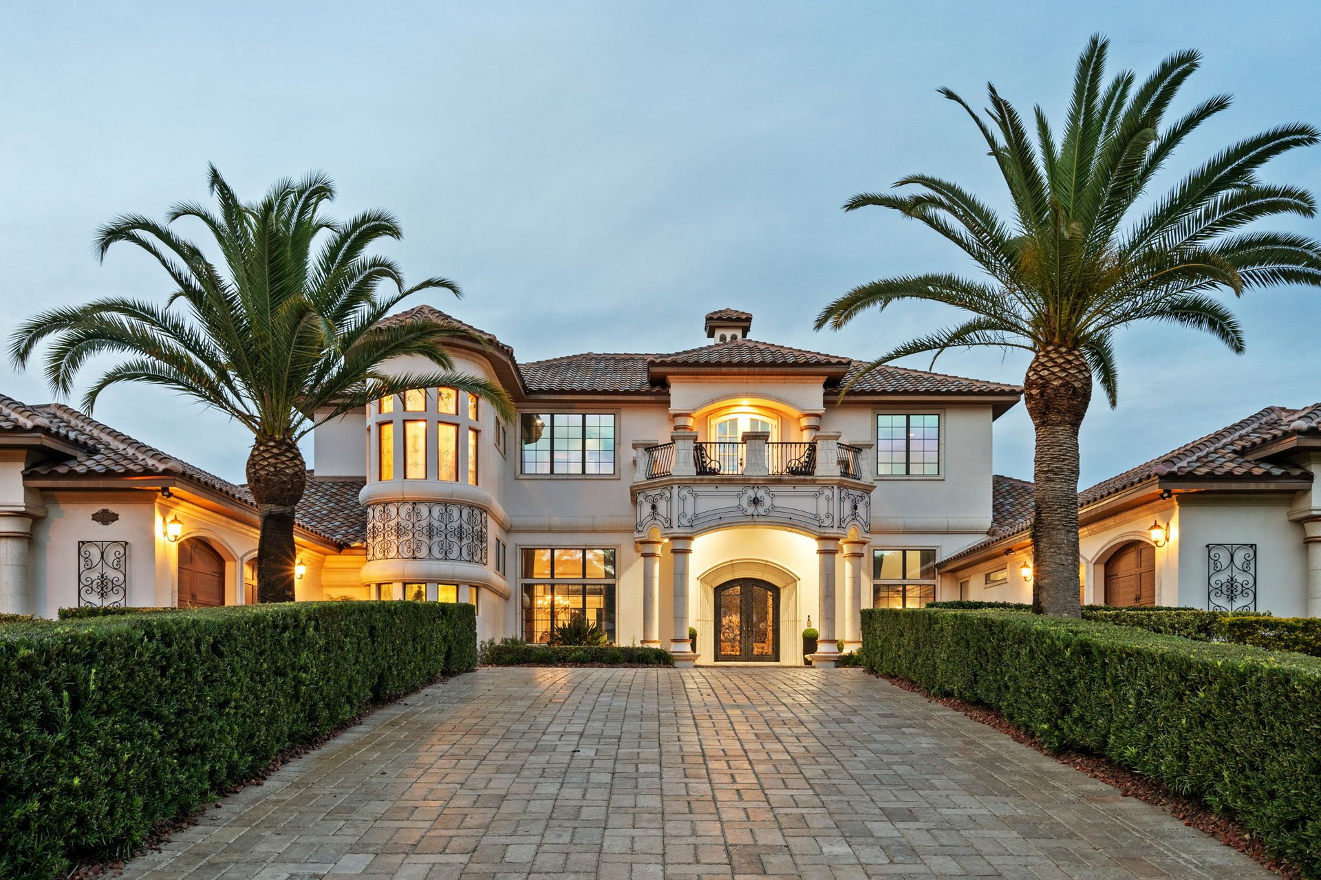 Two-story stucco home with ornate balcony, paved driveway, clipped hedges, and tall palms at dusk.