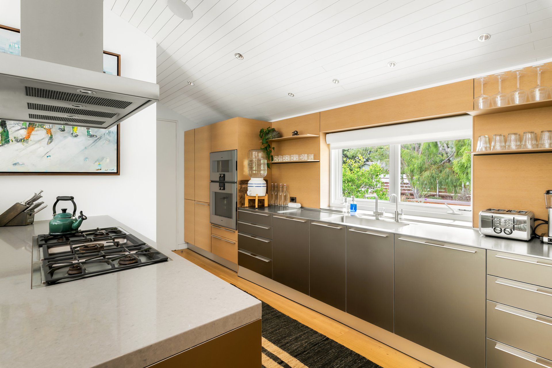 Modern kitchen with wood cabinetry, gas cooktop, and a large window overlooking greenery.