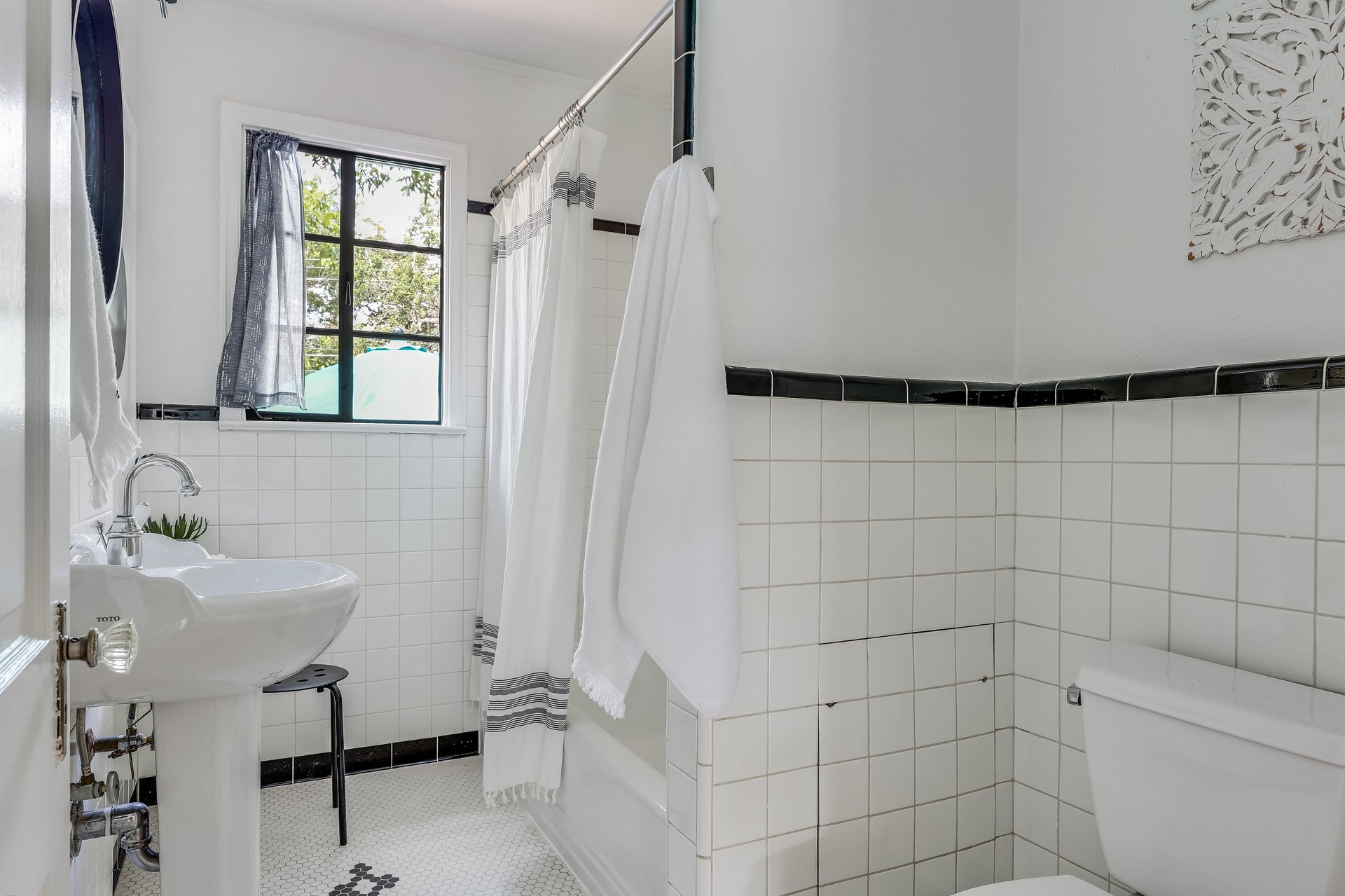 Bright tiled bathroom with a pedestal sink, curtained bathtub, and black-framed window.