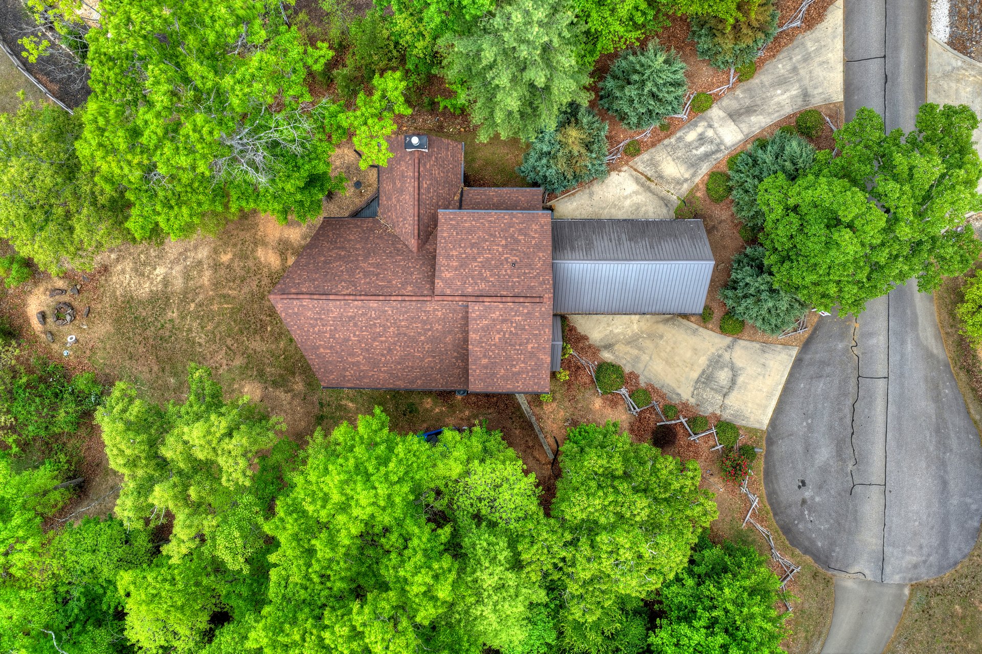 Aerial view of a brown-roofed home surrounded by trees, with a curved driveway and attached metal-roofed structure.