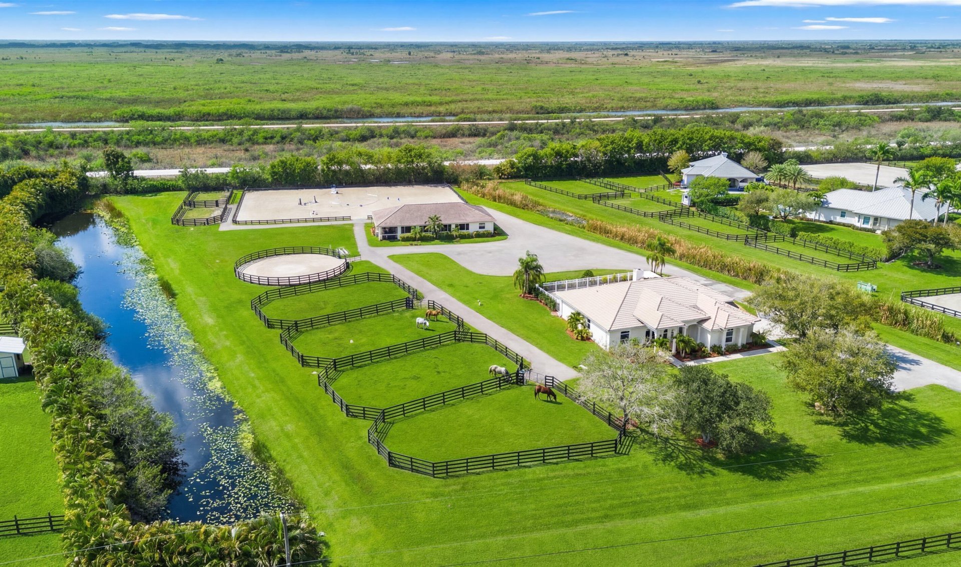 Aerial view of an equestrian estate with fenced paddocks, riding arena, homes, and a canal.