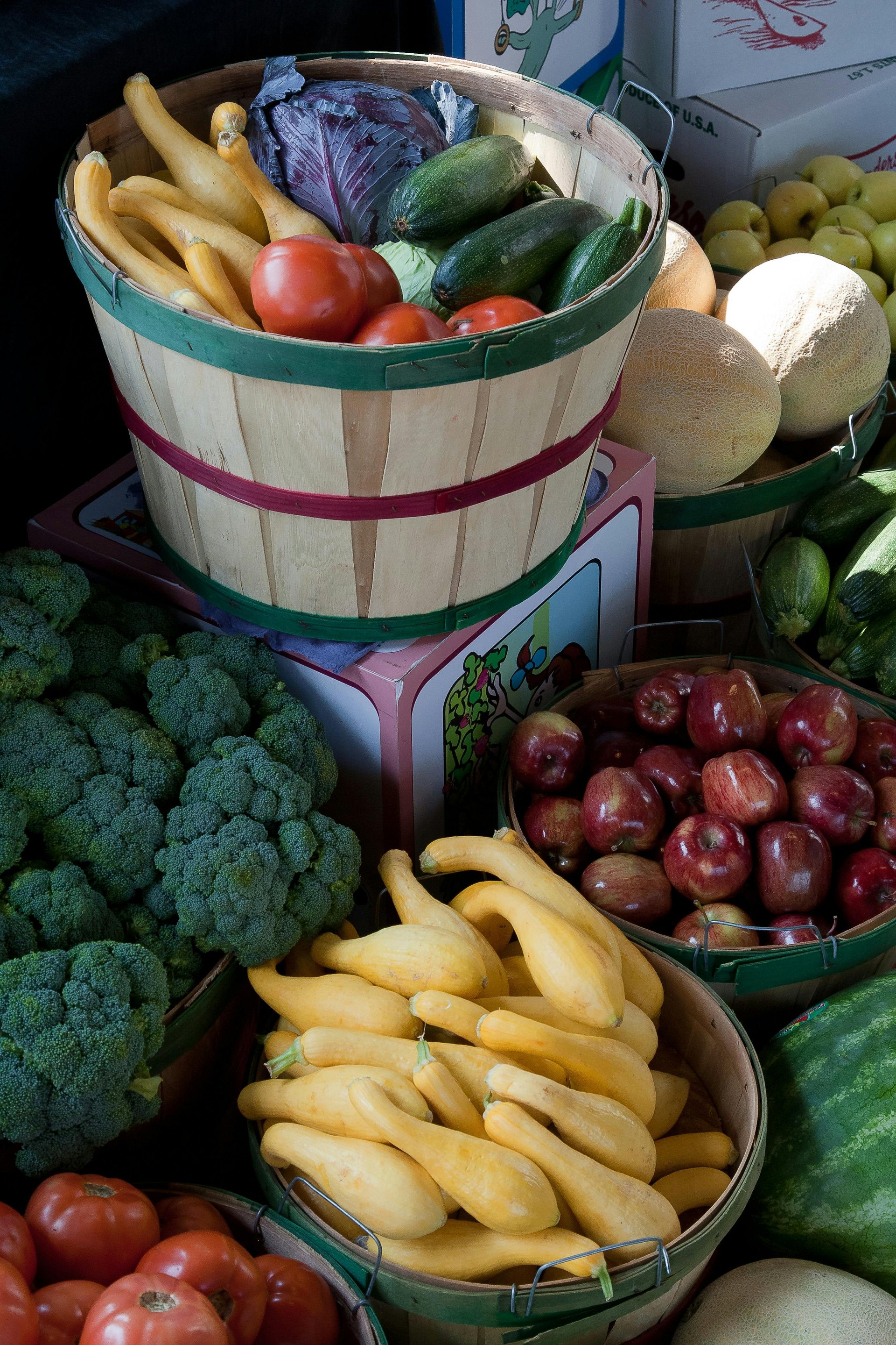 A variety of fresh vegetables and fruits, including squash, tomatoes, broccoli, and apples, displayed in baskets.